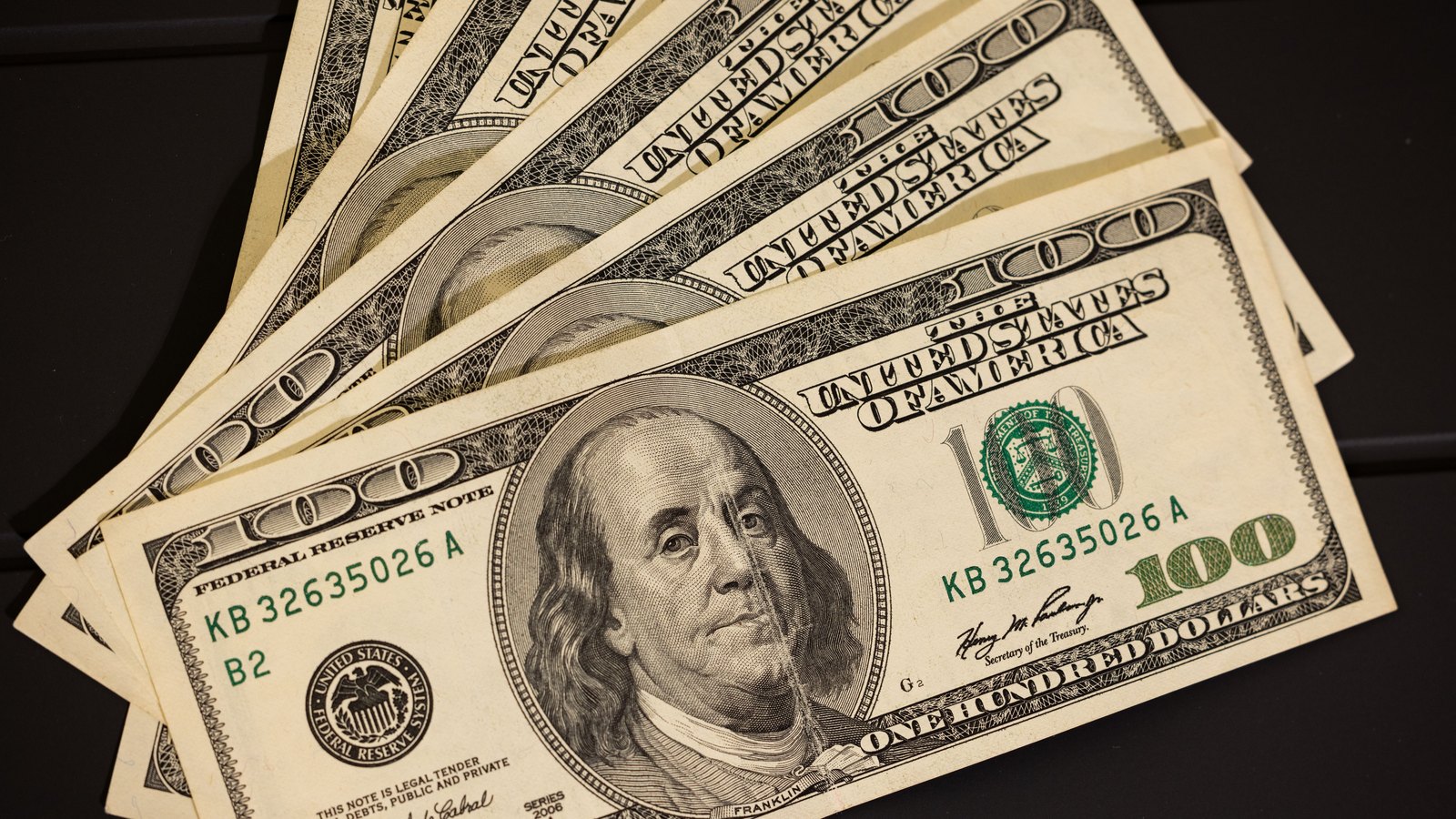 Six United States one hundred dollar bills fanned out flat on a dark surface, the Franklin portrait, the green Treasury seal and the Federal Reserve Note lettering facing the camera.