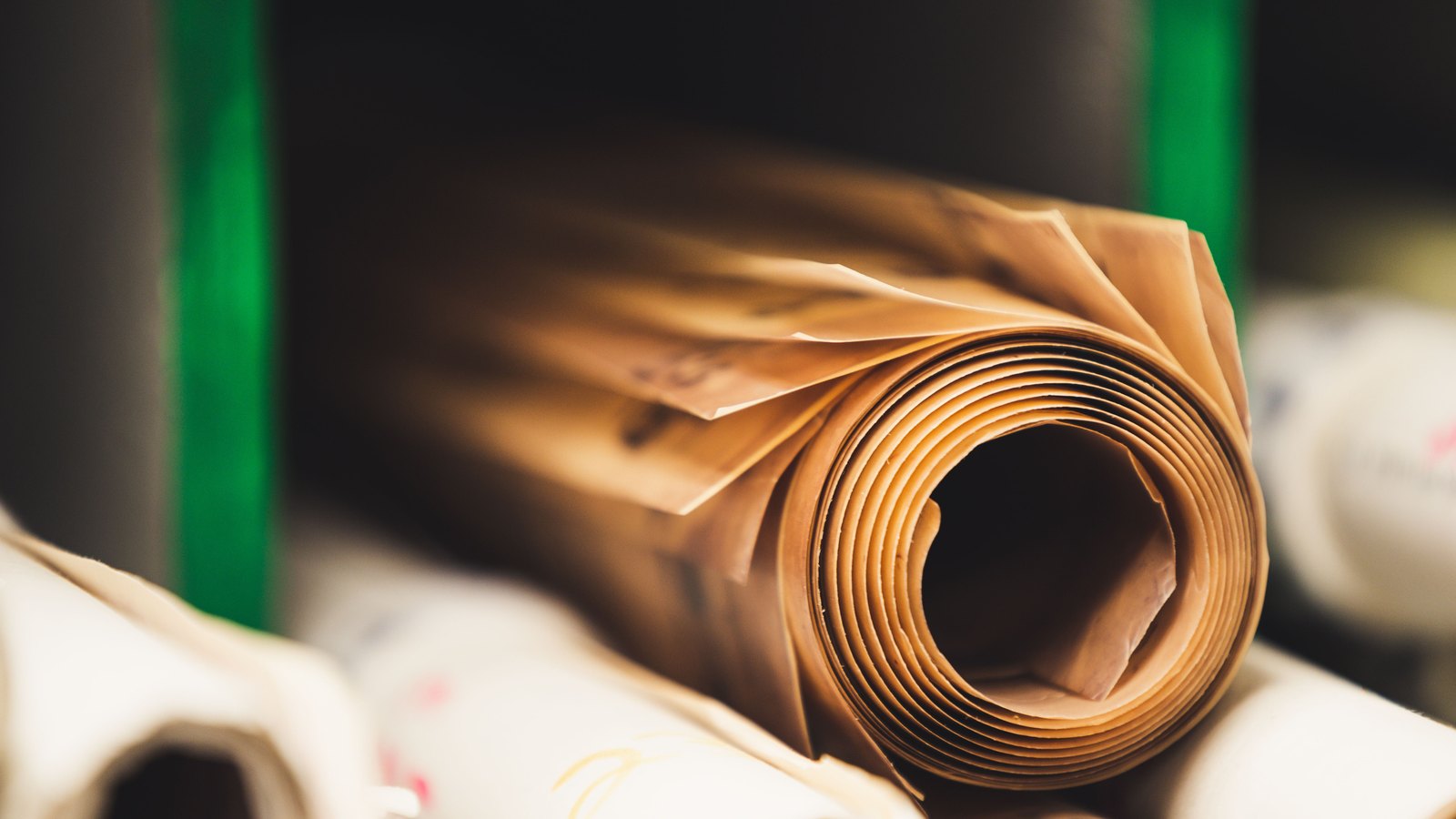 A close view of a roll of brown kraft paper lying on its side, the coiled layers filling the right of the frame with the open core in shadow, and further pale rolls out of focus around it.