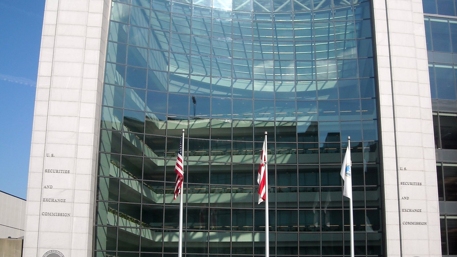The headquarters of the United States Securities and Exchange Commission in Washington, seen straight on: a curved wall of green tinted glass set between two pale stone piers, each pier carrying the words U.S. Securities and Exchange Commission incised in capitals, the top of the Commission seal just visible at the lower left, and three flagpoles carrying the United States flag, the flag of the District of Columbia and the Commission flag standing in front. Nobody is in the frame.