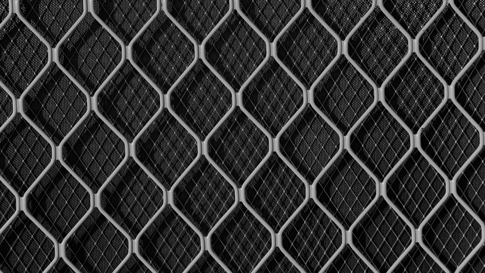 A flat pale metal lattice of repeating diamond shapes laid over a darker woven mesh behind it, filling the frame.