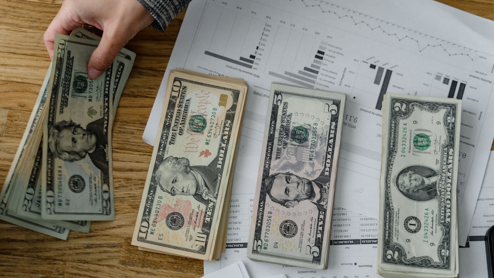 US dollar banknotes arranged over printed financial charts as a hand lifts a stack