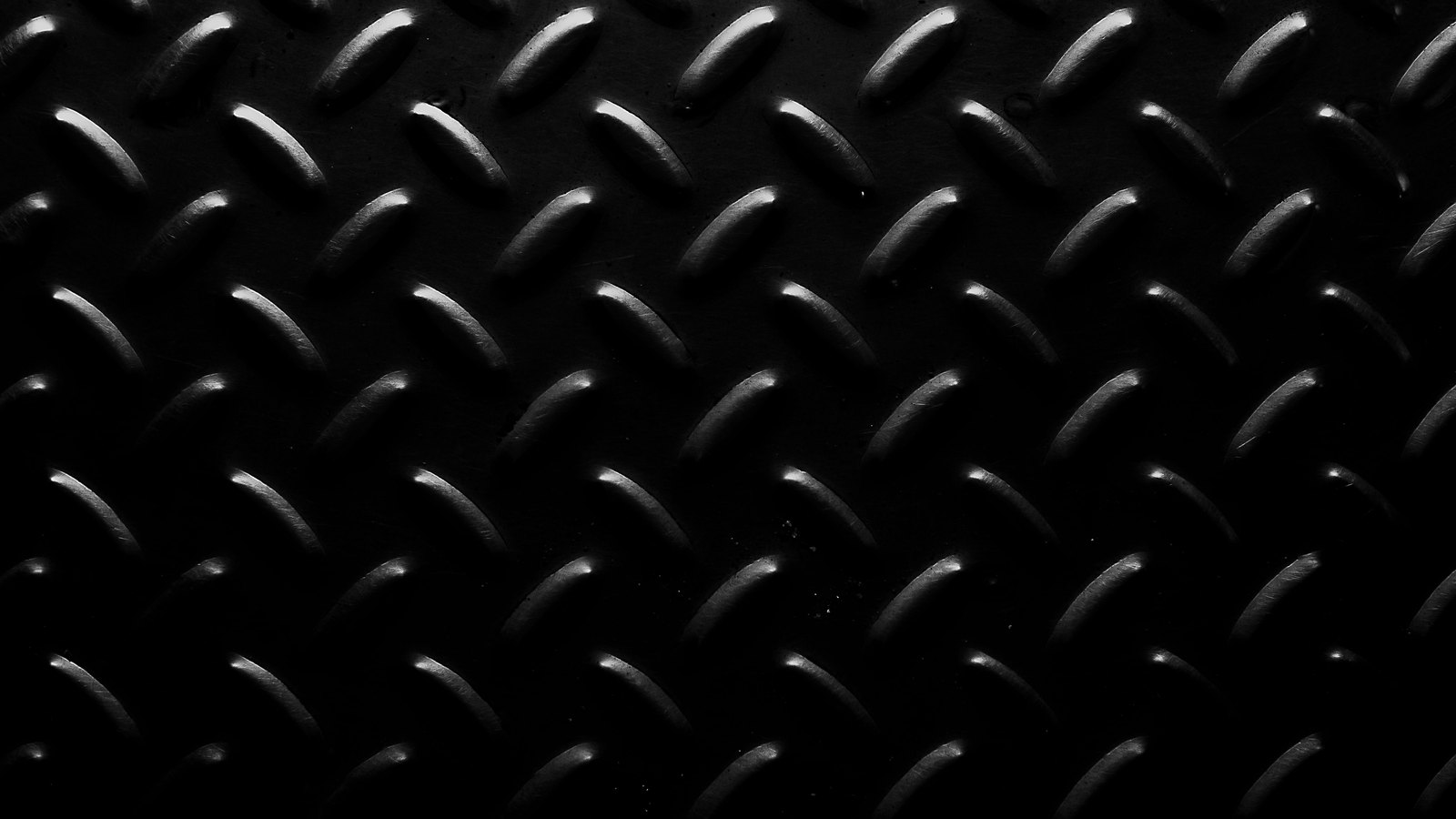 A close photograph of steel tread plate filling the whole frame, rows of raised diamond ribs catching a narrow light across a near black surface that falls into shadow at the corners. No text, brand mark, person or location is in view.