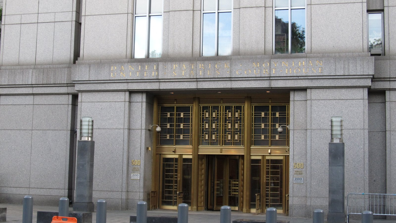The street level entrance of the Daniel Patrick Moynihan United States Courthouse in Manhattan, a grey granite facade with the building's name cut in gold capitals above a bank of tall brass doors, bollards along the pavement in front.