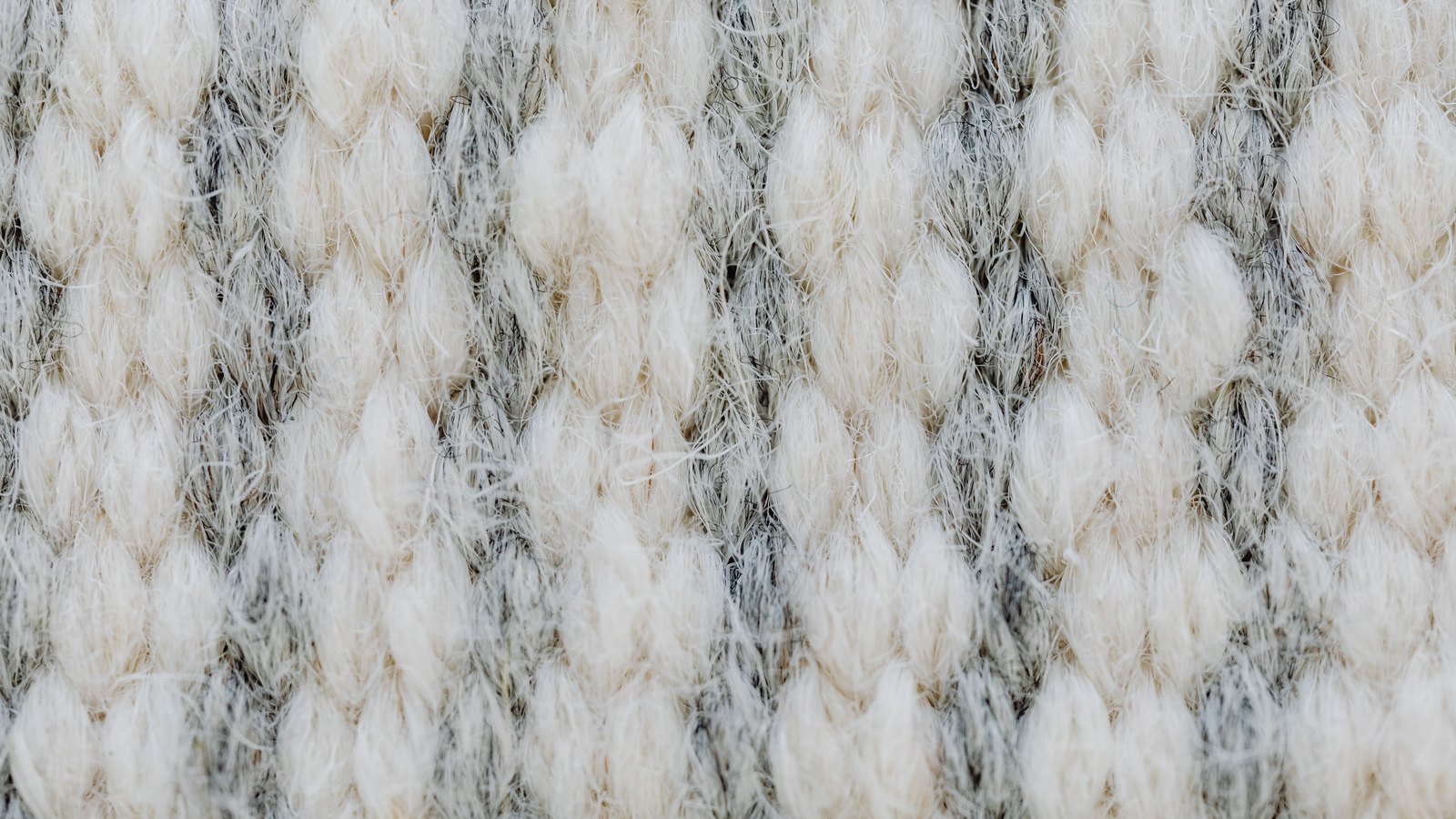 A macro crop of a coarse hand knitted wool in cream and marled grey, the plump loops standing in rows with loose fibres catching the light across the whole frame. No text, brand mark, person or place is in view.