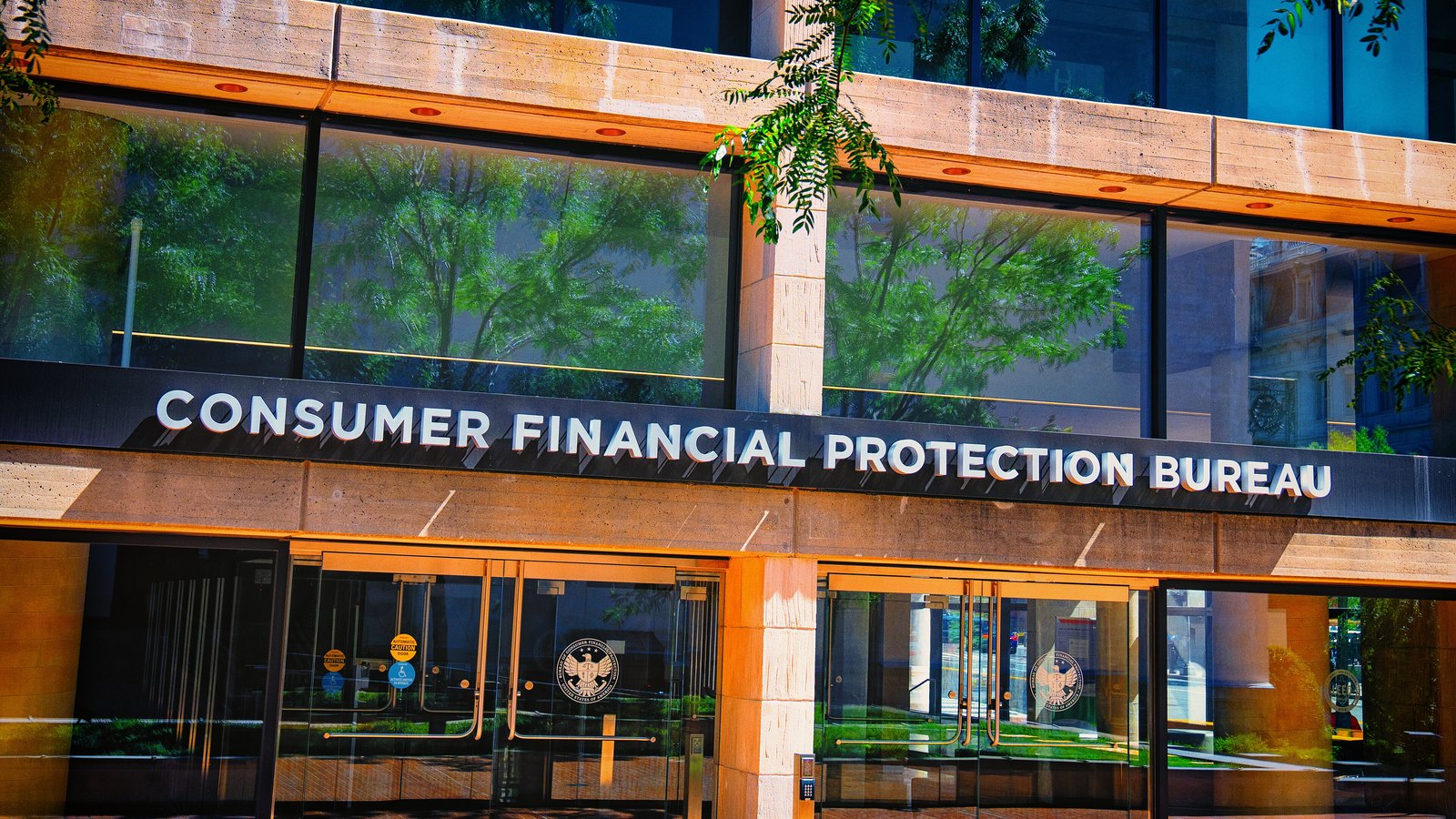The entrance to the Consumer Financial Protection Bureau headquarters in Washington, the agency name in raised metal letters on a dark fascia above glass doors carrying its seal, trees reflected in the windows above.