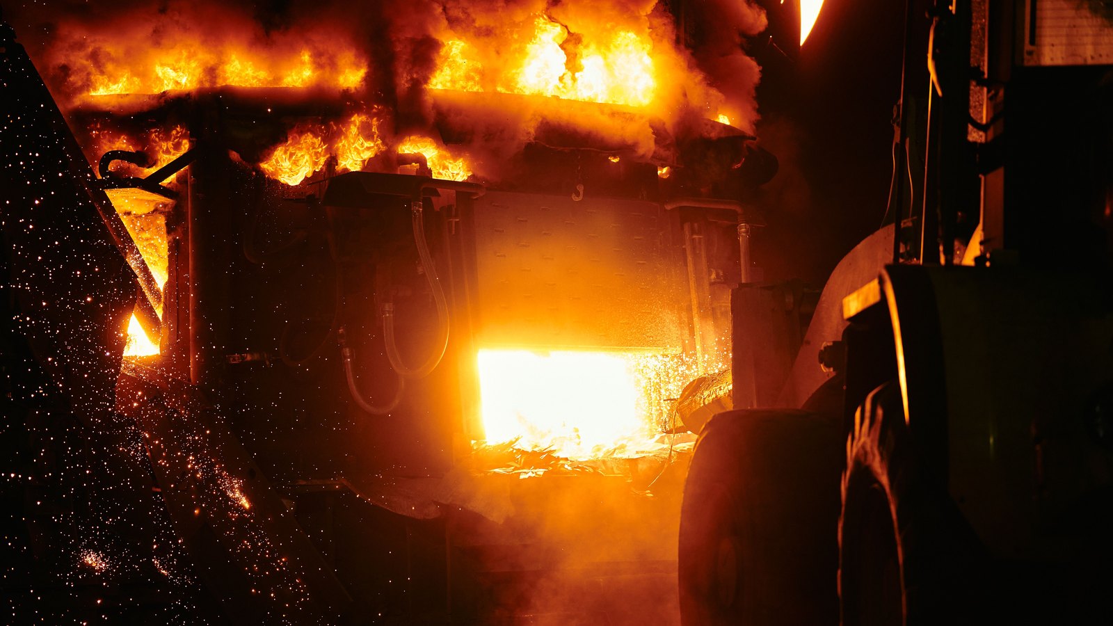 The open mouth of an industrial furnace at night, flame and smoke rolling from the top of the vessel and sparks falling in front of it, with the dark silhouette of a wheeled loader at the right of the frame. No people, lettering or brand marks are in view.