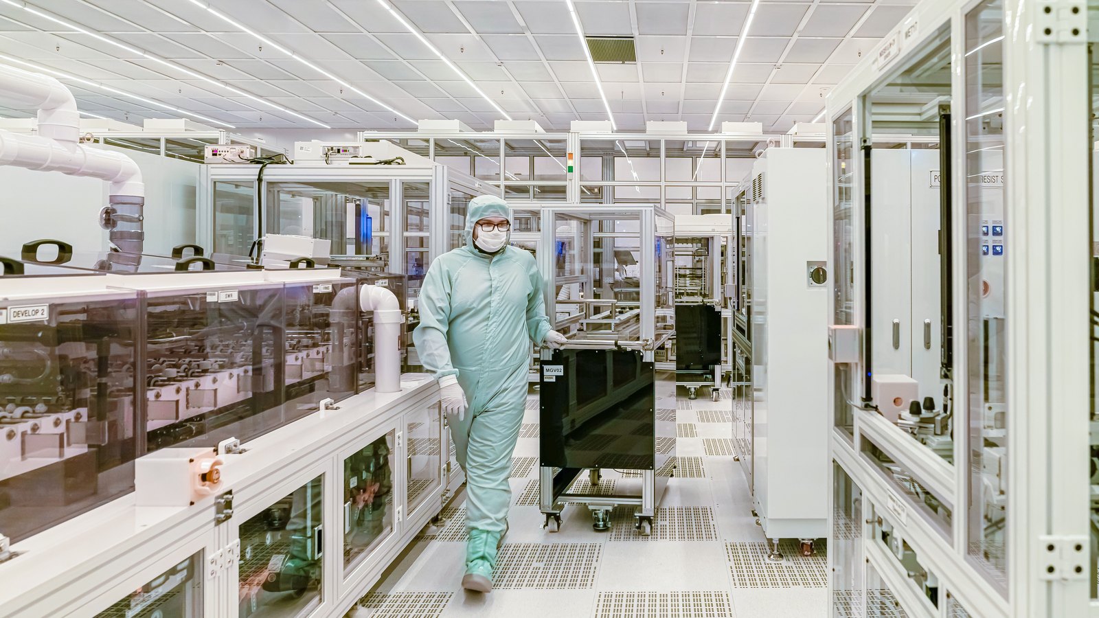 A wide aisle in a semiconductor clean room, lined on both sides with white and glass process tool cabinets under bright ceiling strip lights. One worker in a pale mint hooded coverall, mask, glasses and white gloves walks toward the camera pushing a low black wheeled cart. Small station labels are on the equipment. No brand, logo, company name or place is visible.