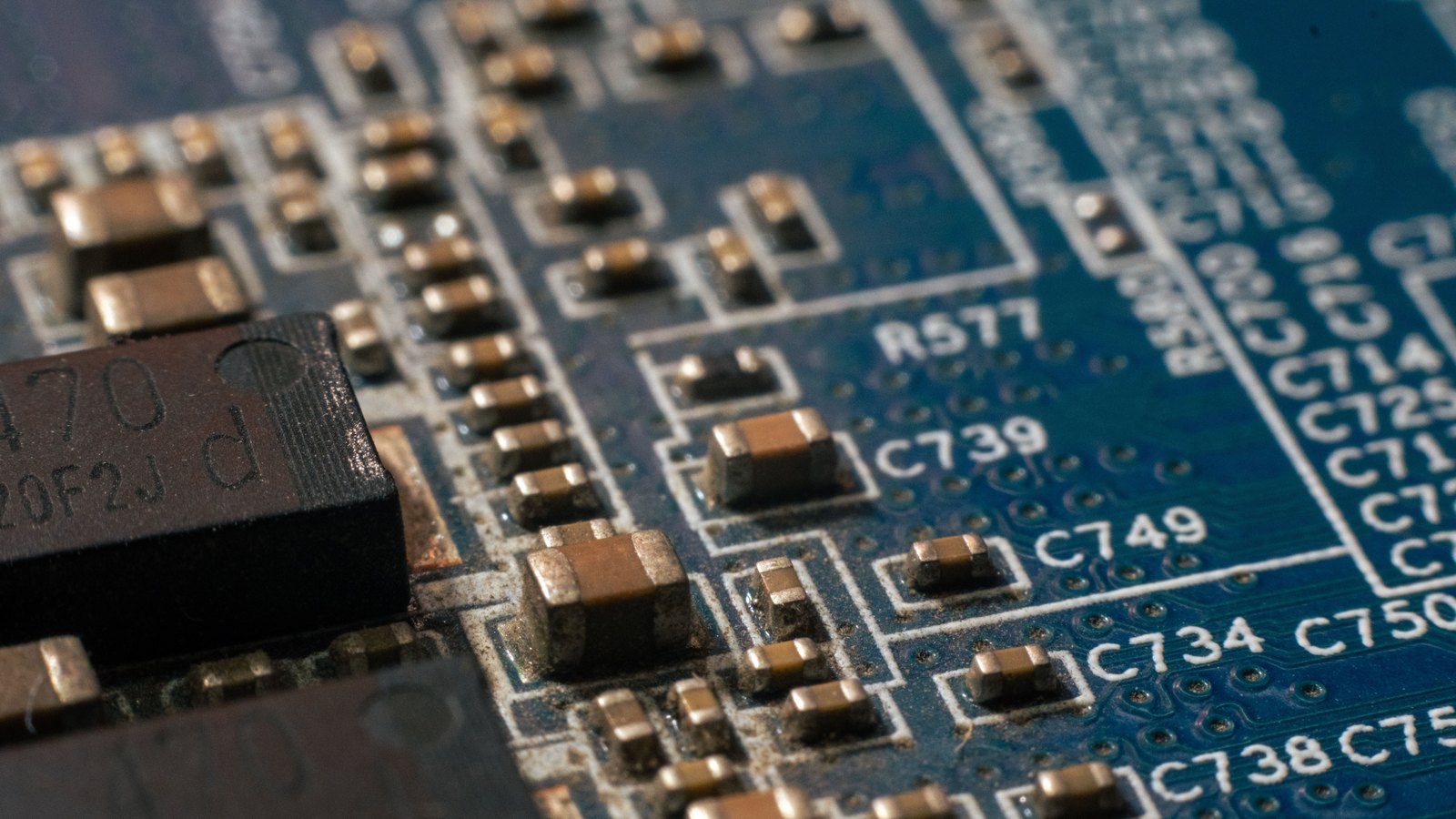 A close macro of a blue printed circuit board at an angle, showing rows of small gold-capped surface-mount capacitors, a black component, and white silkscreen part codes reading C739, C749, R577 and C734.