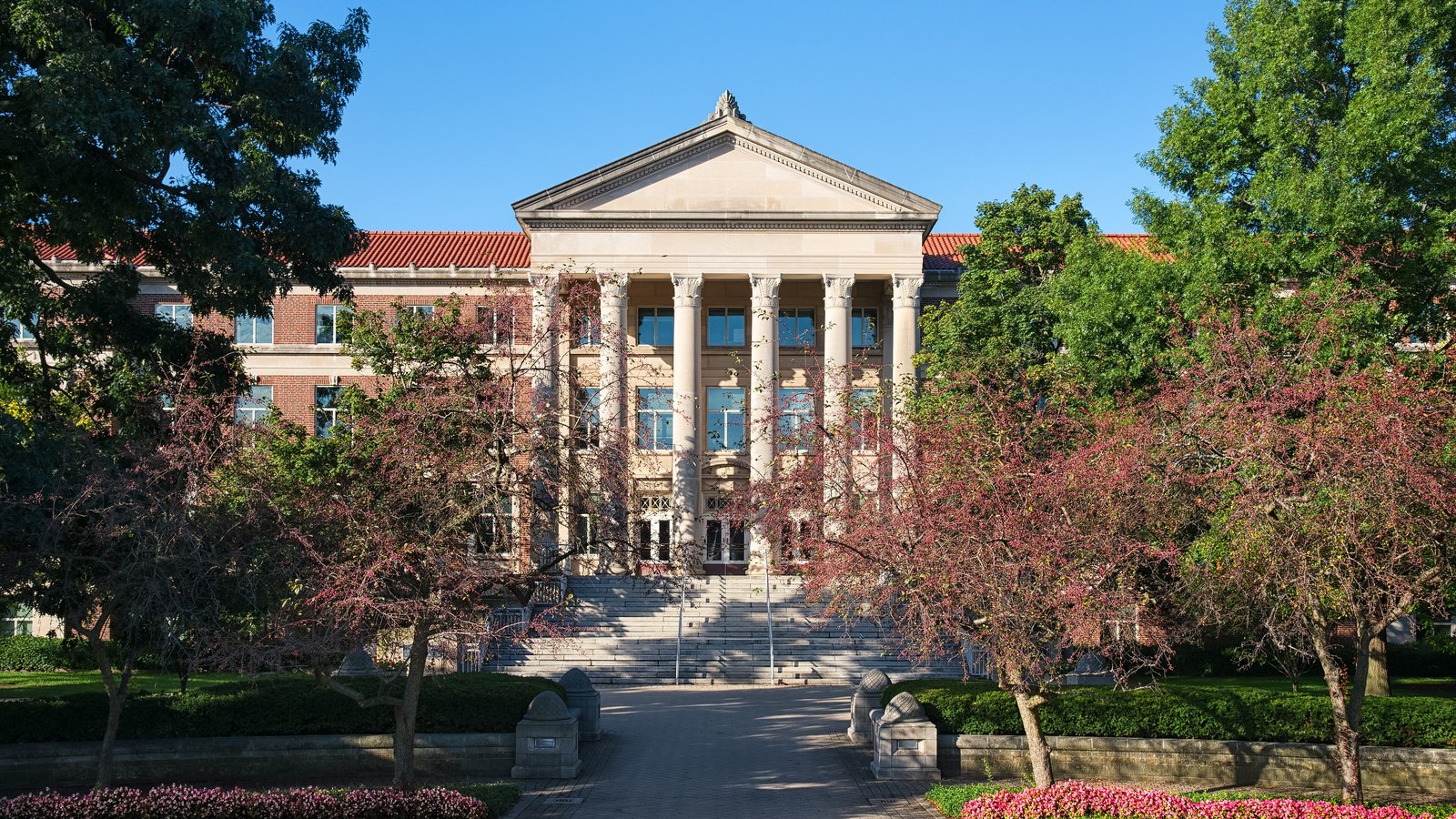 A three storey red brick building with a projecting limestone portico of six tall fluted columns under a plain triangular pediment, photographed straight on from the end of a brick walk. Flowering crabapple trees in pink blossom stand on both sides, a broad flight of stone steps rises to the doors, and clipped hedges with beds of pink and white flowers run across the foreground under a clear blue sky. No people, signs or lettering are in the frame. Hovde Hall of Administration at Purdue University in West Lafayette, Indiana, photographed in 2016.