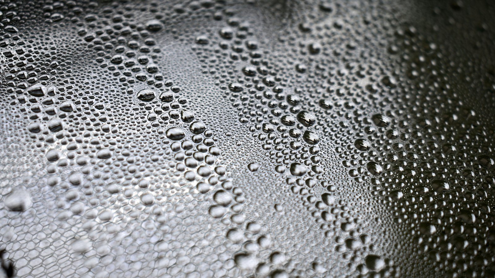 A close photograph of condensation on a grey surface, thousands of clear water droplets of many different sizes covering the whole frame, with a soft band of highlight running across the middle and the corners falling darker. It is used as a generic illustration. No text, brand mark, person, building or place is in view.