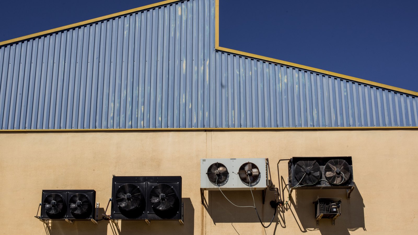 Four banks of industrial cooling fans mounted along the plain exterior wall of a warehouse beneath a blue metal roof.