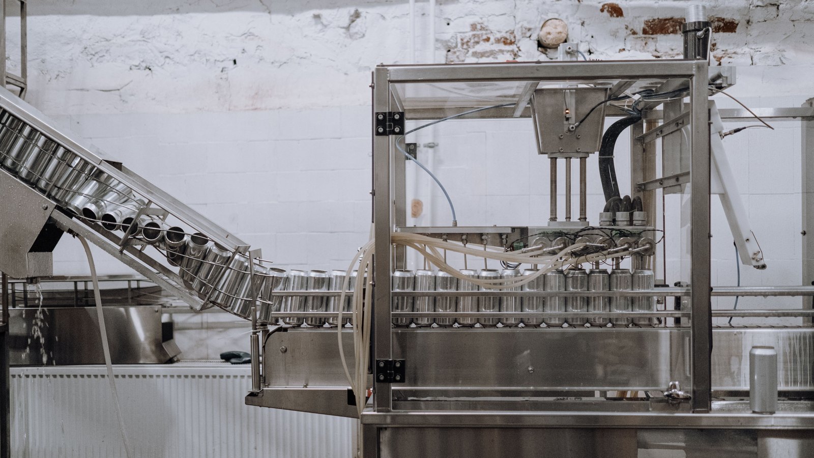 Plain unlabelled aluminium cans travelling along a stainless steel conveyor into a filling machine, with an inclined feed track carrying more cans down from the left and a painted brick wall behind. No person, brand, lettering or place is in the frame. An illustrative photograph.