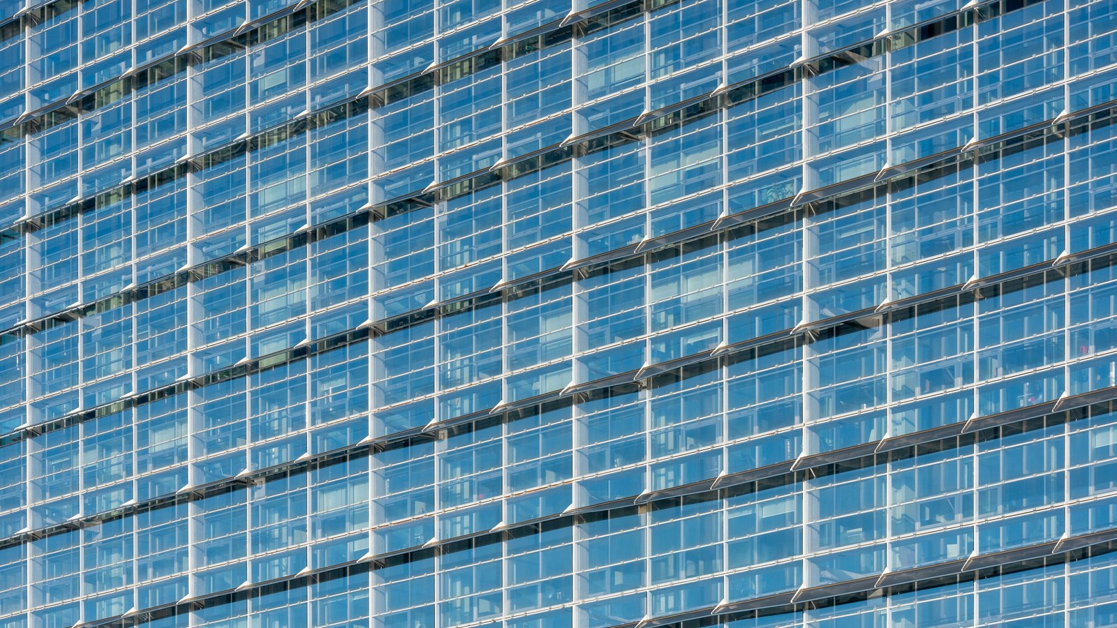 A close crop of a modern office tower curtain wall, filling the frame with a repeating grid of blue tinted glazing panels, pale metal mullions and horizontal shading fins set at a slight diagonal. No text, signage, people or sky are in the frame.