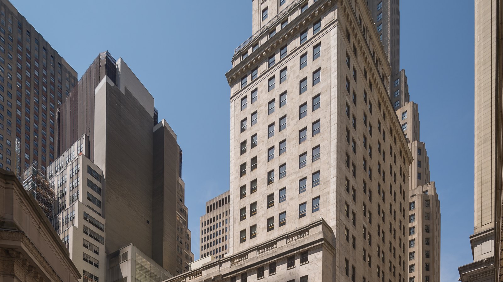 Office towers in the Wall Street financial district in Manhattan, looking upward from street level: a pale stone tower with a stepped setback fills the right of the frame, darker slab-sided blocks stand behind it to the left, and the sky above is clear. No people, signage or lettering are visible.