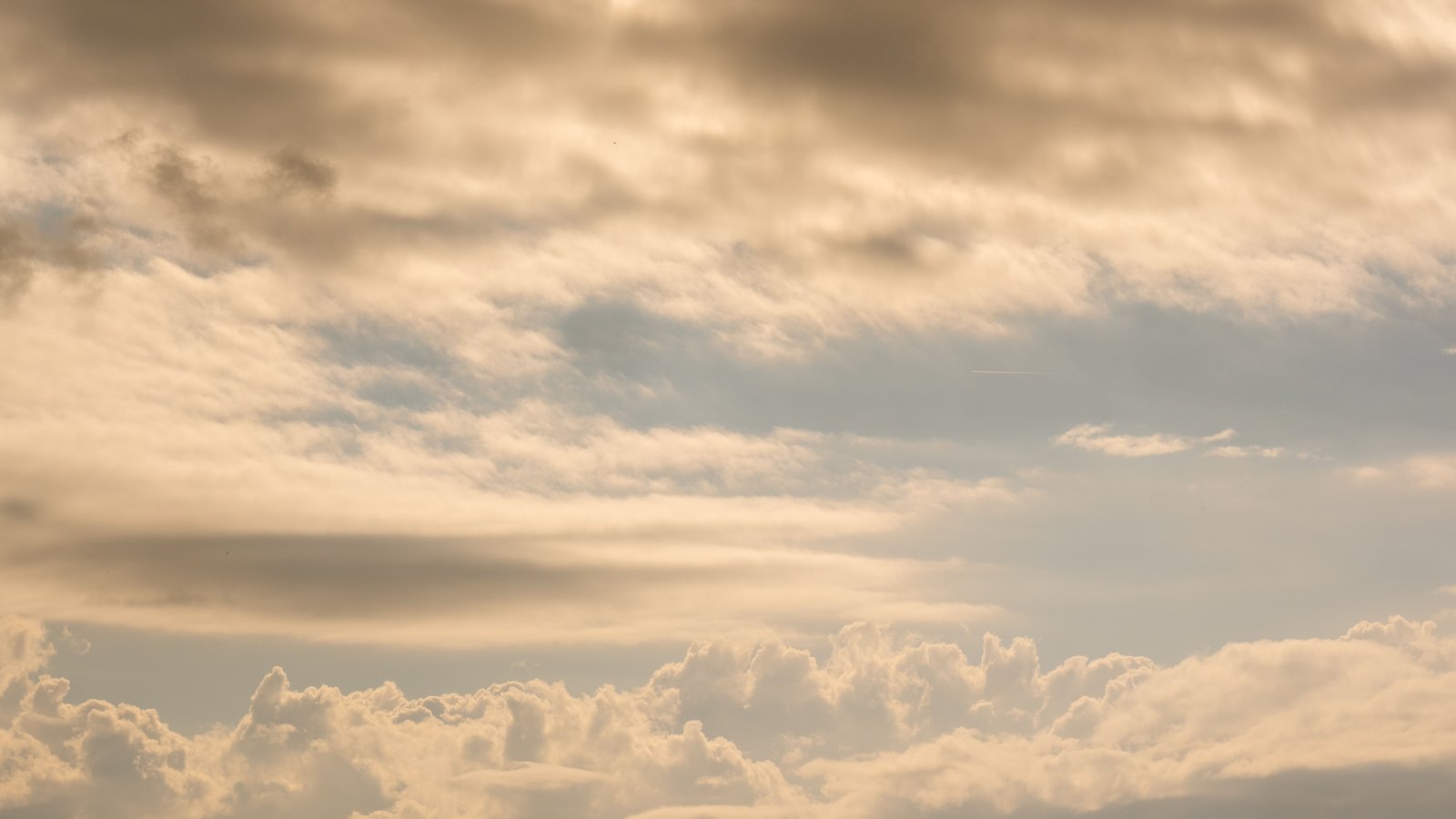 An open sky filled with soft cream and grey cloud lit warm by low sun, banded cloud across the upper frame and a rise of cumulus along the bottom. No horizon line, no ground, no buildings, no people, no vehicles, no signage and no lettering of any kind.