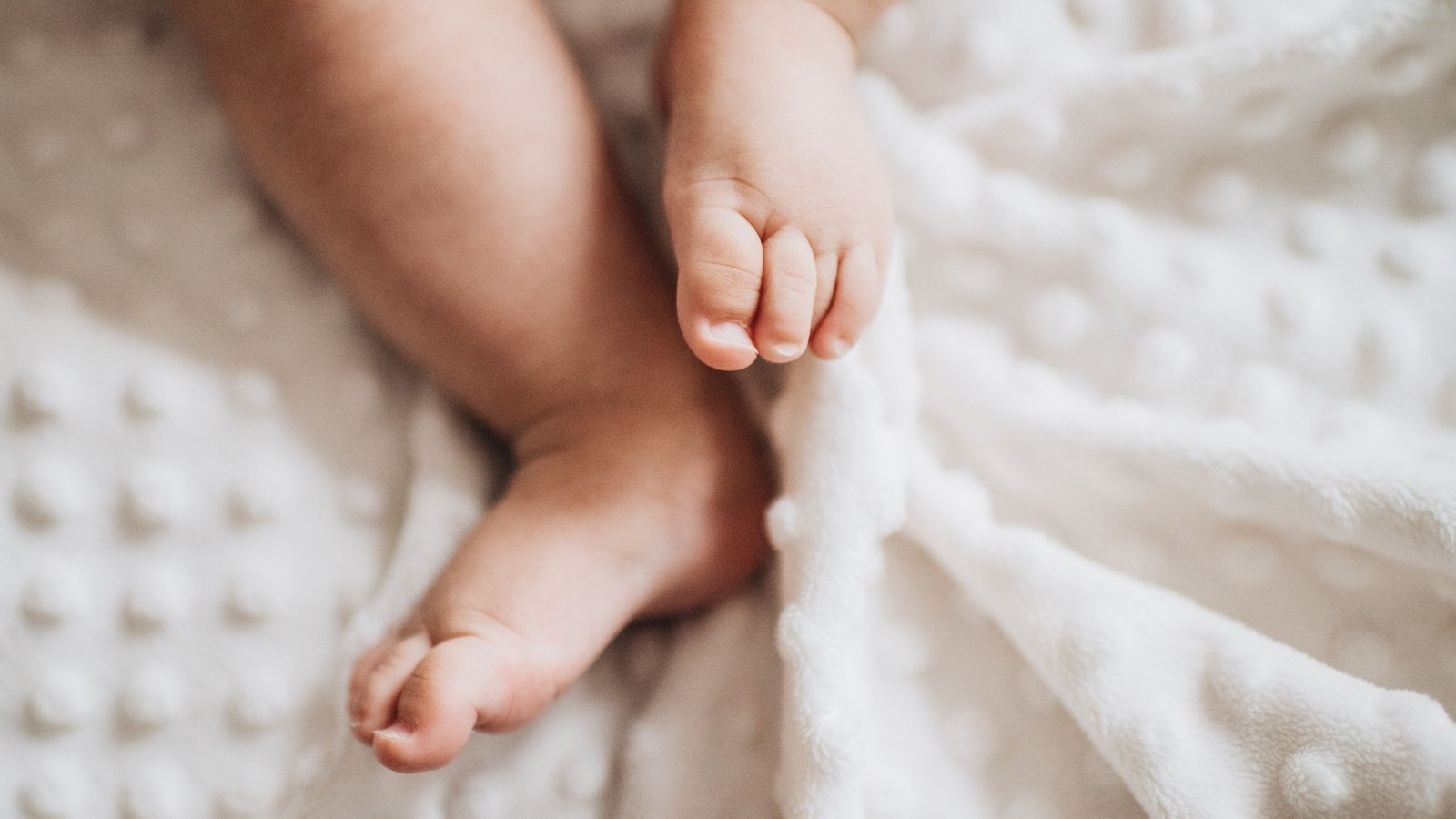 A newborn's bare feet rest on a white knitted blanket in close-up.