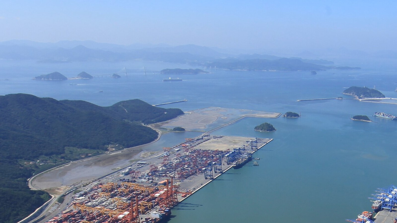 An aerial view of container terminals lining a broad shipping channel at Busan, with stacked containers, gantry cranes, wooded hills and islands in a hazy sea beyond.