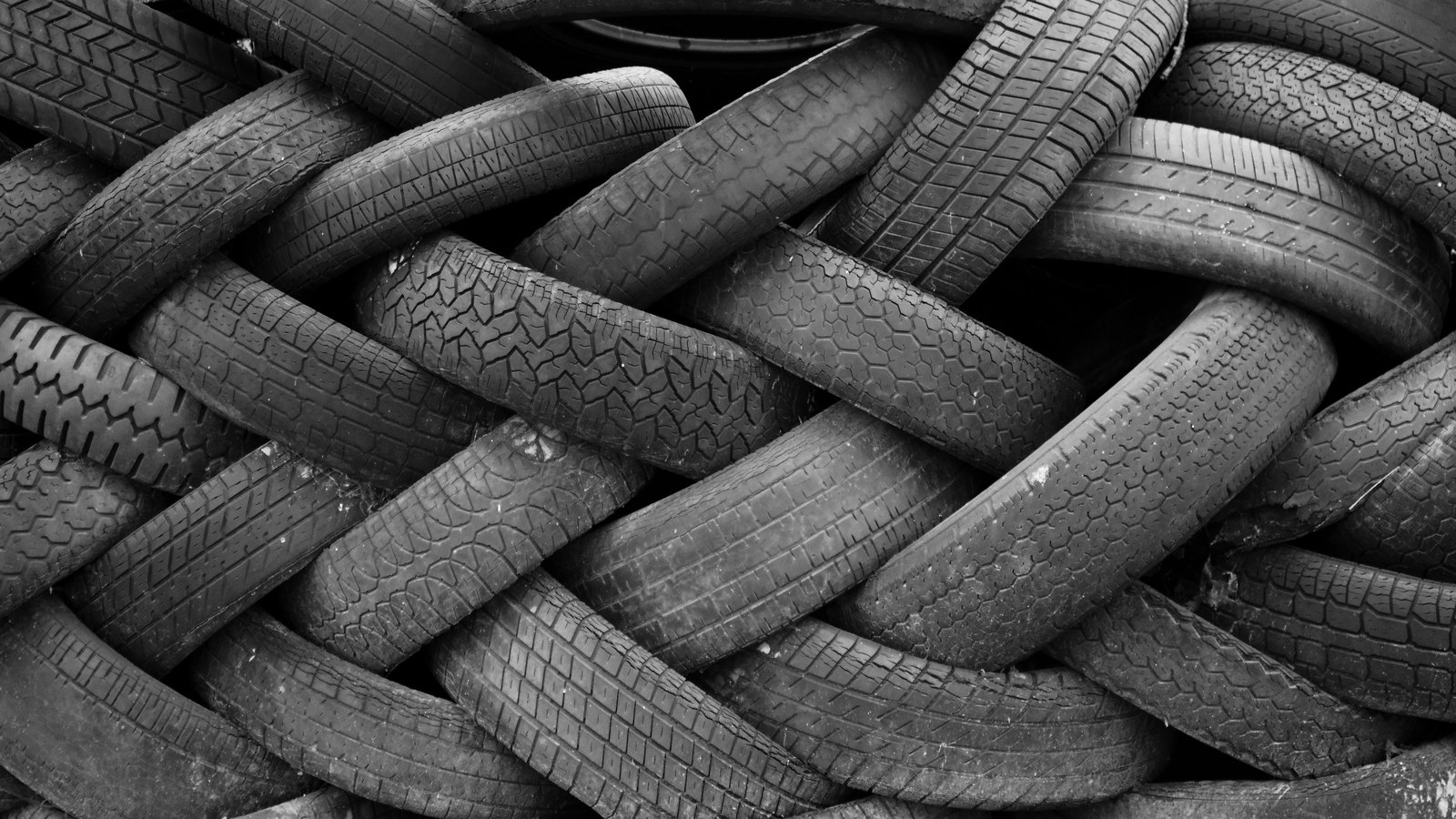 A black and white photograph of a dense stack of worn tires lying on their sides and overlapping, filling the frame with bands of tread pattern in several different designs. No aircraft, people, buildings, signage or lettering is in the frame. An illustration of tire rubber and tread, not a photograph of this aircraft or its landing gear.