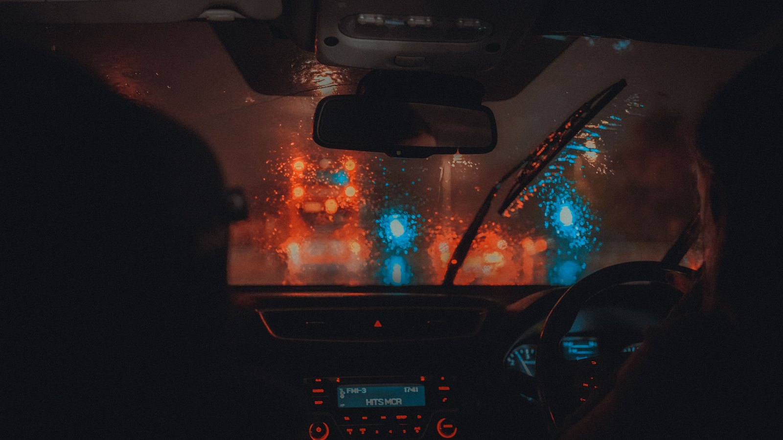 Rain covers a windshield as traffic lights blur beyond dark vehicle occupants, not a photograph of this advisory.