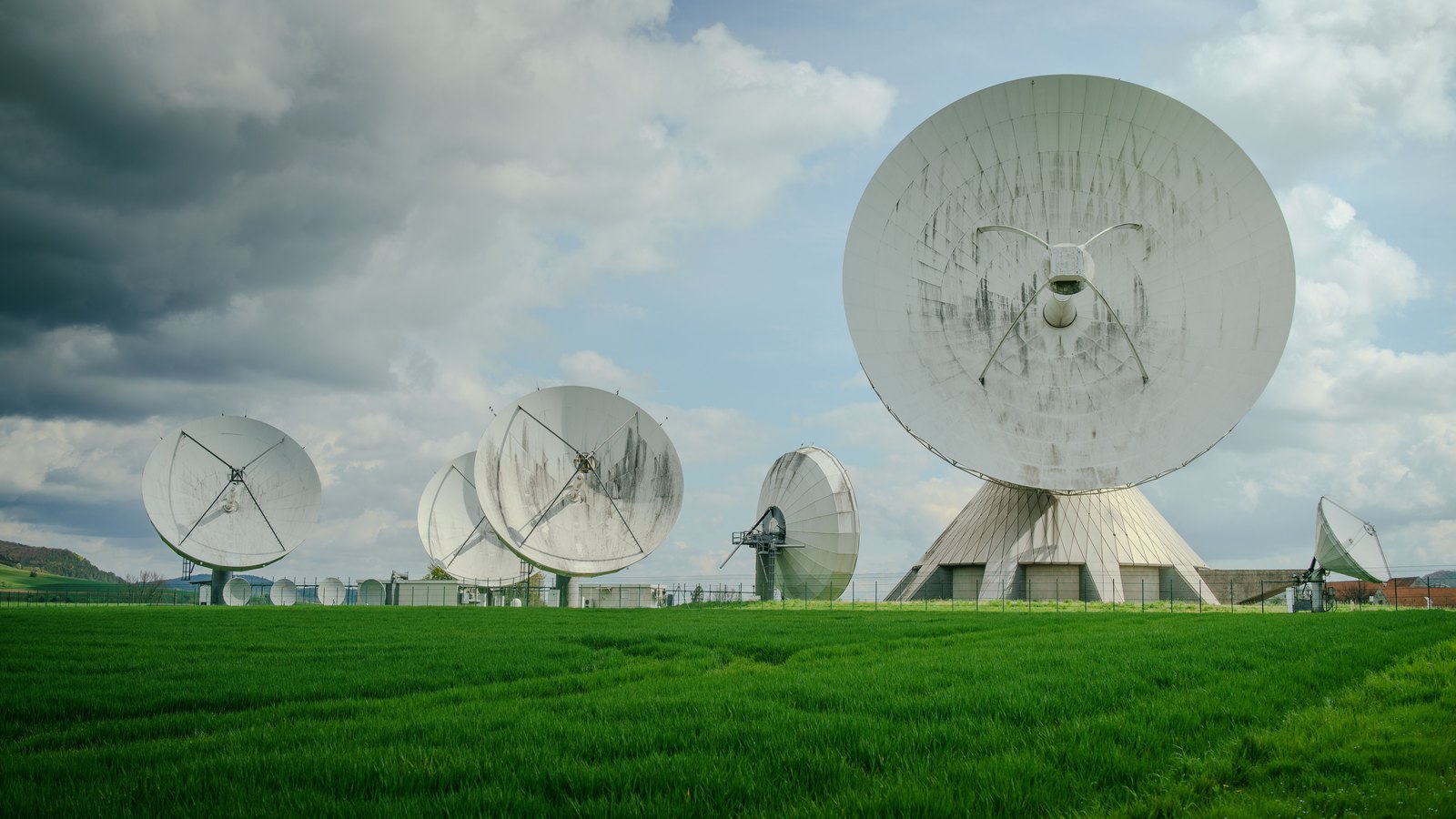 Five large white parabolic dish antennas of different sizes standing in a field of tall green grass, angled upward, under a sky of heavy grey and white cloud with patches of blue. A low fence and small plain outbuildings run behind them. No text, sign, logo, vehicle or person is visible.