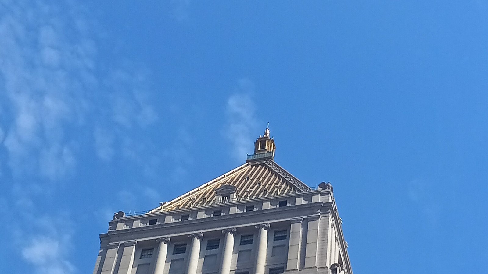 The top of the Thurgood Marshall United States Courthouse in New York seen from street level against a clear blue sky, showing the steeply pitched gilded pyramid roof, the small gold lantern and spire at its apex, and the colonnaded upper storeys below the cornice. No people, signage or lettering are in view.