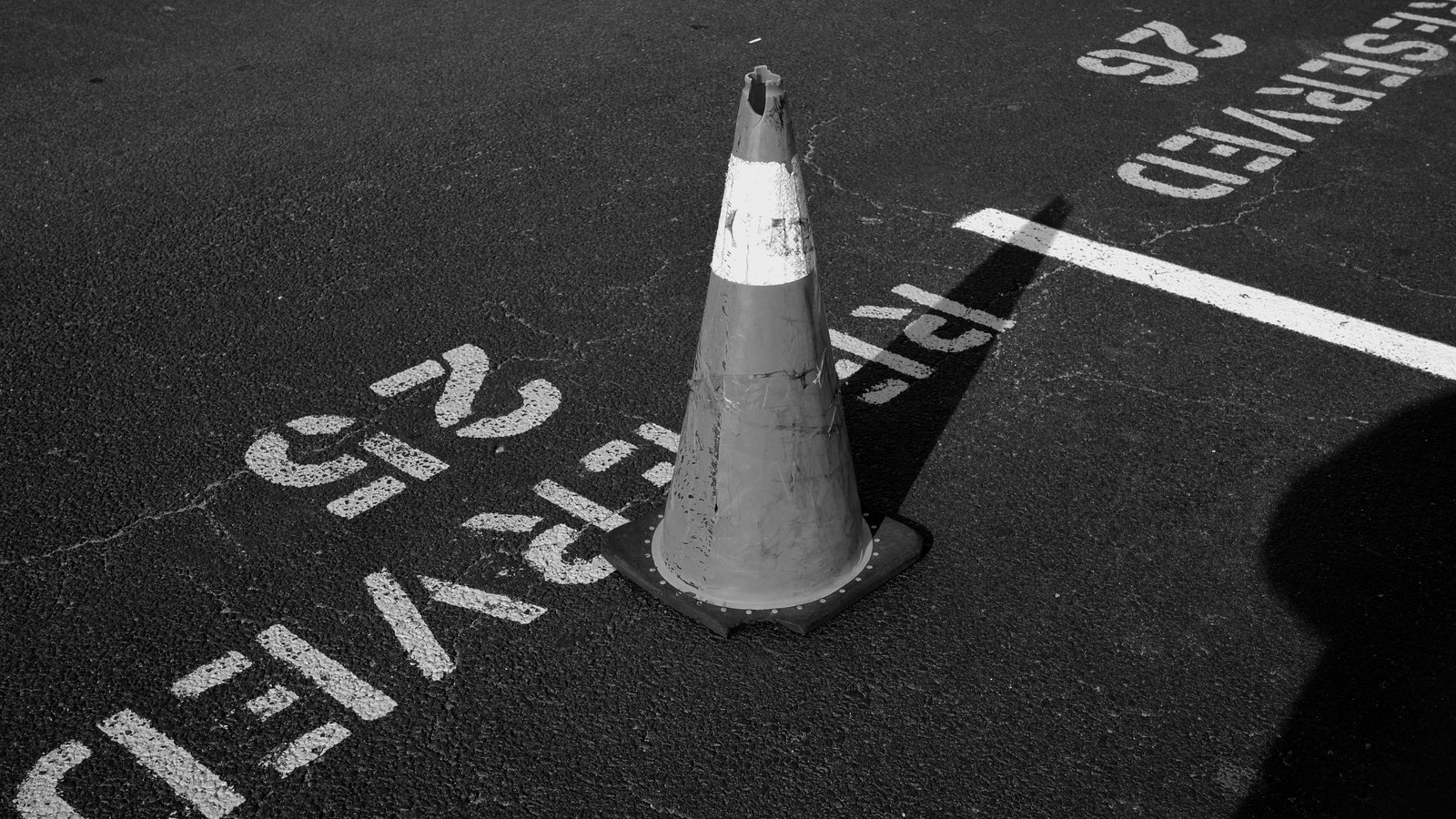 A black and white photograph of one scuffed traffic cone standing on cracked asphalt, its reflective band catching the light, with painted parking bay lines and the stencilled word reserved running upside down across the surface beside it. No people, vehicles, road signs, place names or brand marks are visible and no location can be identified from it. A generic illustration of traffic control, not a photograph of the Stan Gober Memorial Bridge, of Goodland or of the work described here.