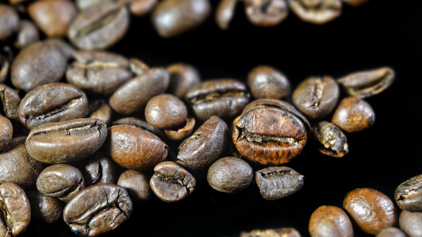 A close photograph of loose roasted coffee beans scattered on a black surface, their split centre seams and oily surfaces visible, with no packaging, label or text in view.