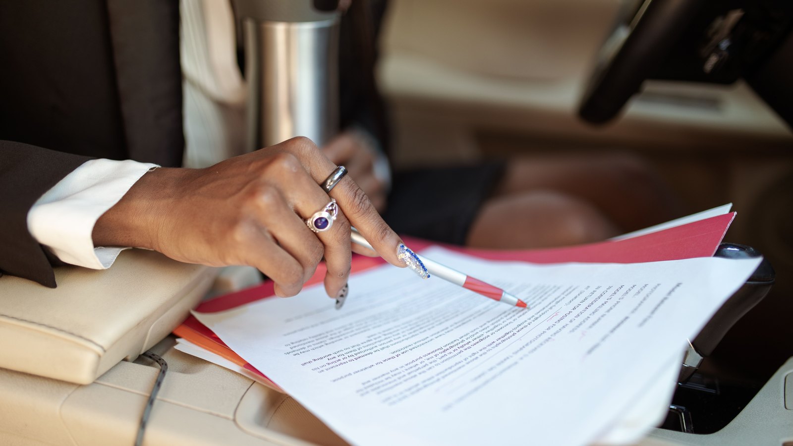 A close view of a hand resting a red and white pen on a loose sheaf of printed paperwork balanced across the front seats of a car, the cream leather upholstery, the edge of the steering wheel and a metal travel cup visible around it, no face in frame and no legible text on the pages. Stock photo, not a photograph of this event.