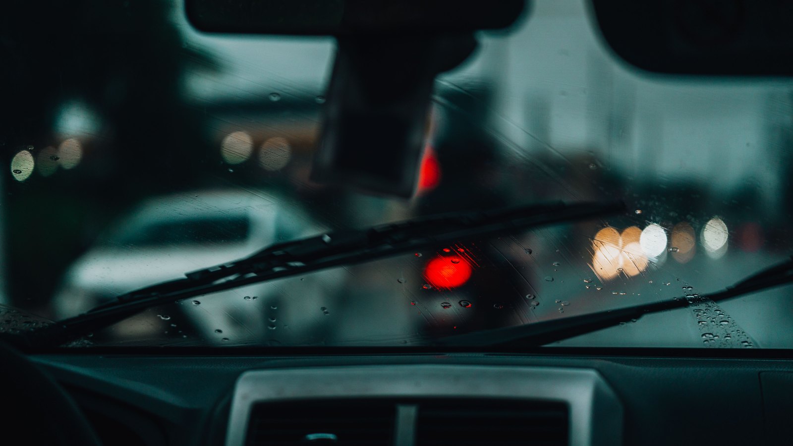 View forward from inside a car through a windscreen streaked with rain, two wiper blades resting across the glass, a blurred red traffic signal and several out of focus warm white and amber lights ahead, and the top of a dark dashboard across the bottom of the frame, with no people, number plates, signs, text or brand marks legible anywhere. A generic illustration, not a photograph of this event.