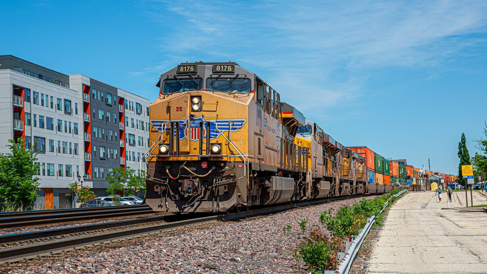 A Union Pacific freight train on straight track in bright sun, led by a yellow and grey locomotive numbered 8176 carrying the railroad's shield emblem. Behind it a second unit and then a long line of double stacked shipping containers in red, orange and green recede toward the horizon. Ballast and a low apartment block line the left of the frame and a paved path runs along the right.