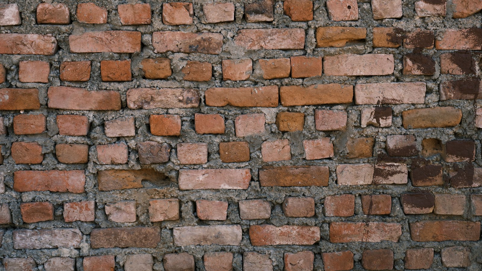 An old brick wall photographed square on and filling the frame. Courses of red, buff and brown bricks of uneven length sit in thick grey mortar that has been pressed roughly into the joints and left proud in places. No paint, lettering or markings are visible.