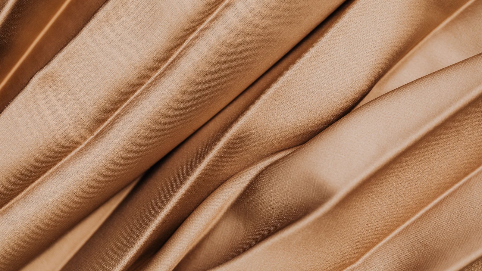A close photograph of pleated tan cloth filling the frame, the folds running diagonally from lower left to upper right, each ridge catching a soft sheen and falling into shadow between, with the weave of the fabric visible across the flat panels. No text, lettering, brand mark, person or place is in view.