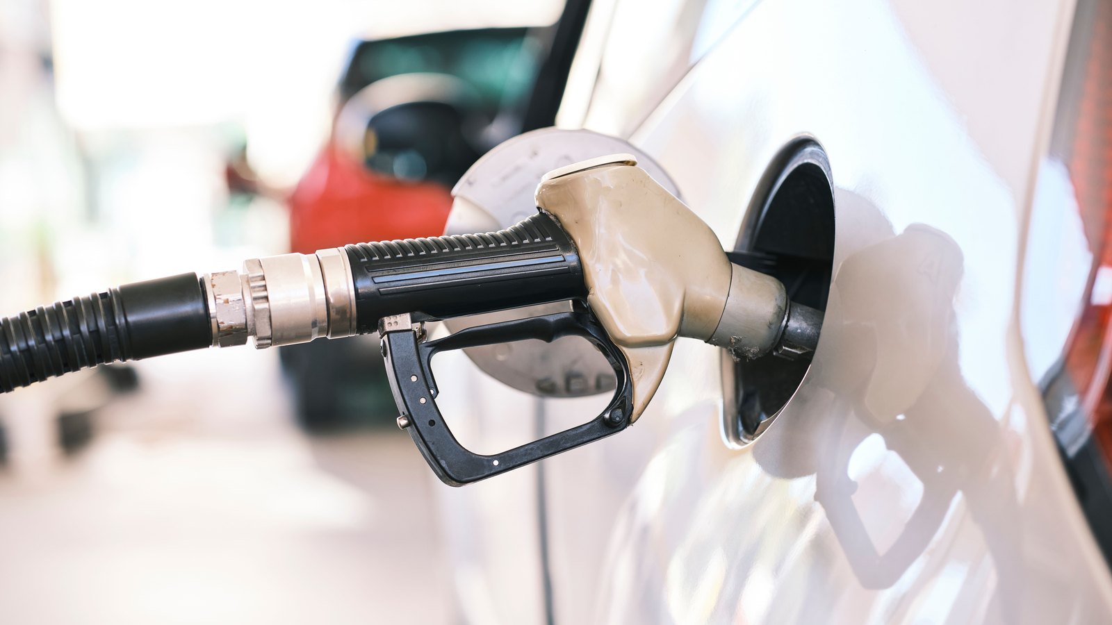 A fuel nozzle seated in the open filler neck of a white car at a service station, the pump hose running out of frame to the left.