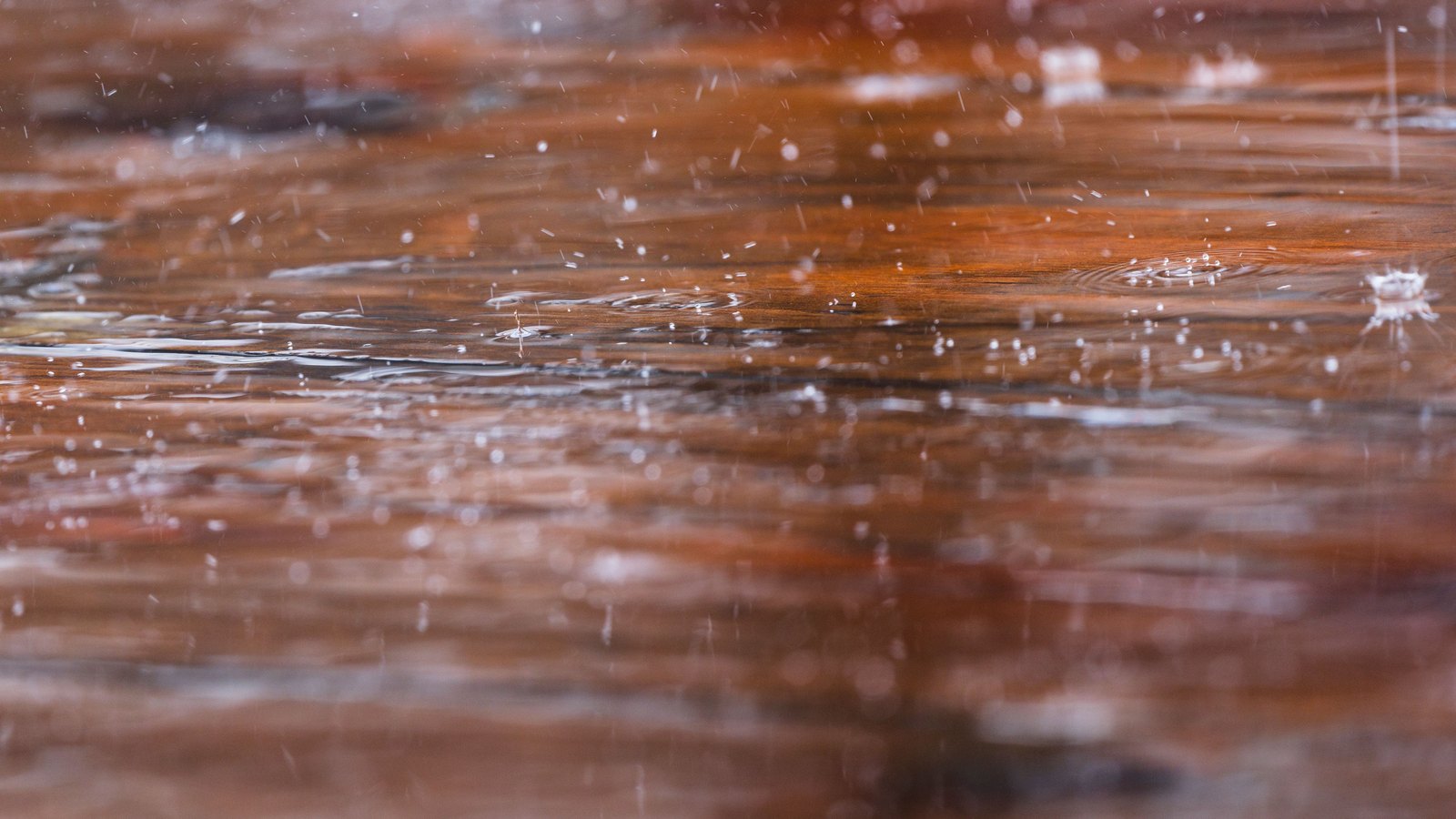 A close macro photograph of rain falling onto a shallow film of standing water lying over a reddish brown wooden surface, with concentric impact rings, small splash crowns and airborne droplets caught in shallow focus. No people, buildings, signs or text are in view. A generic illustration, not a photograph of Collier or Lee County.