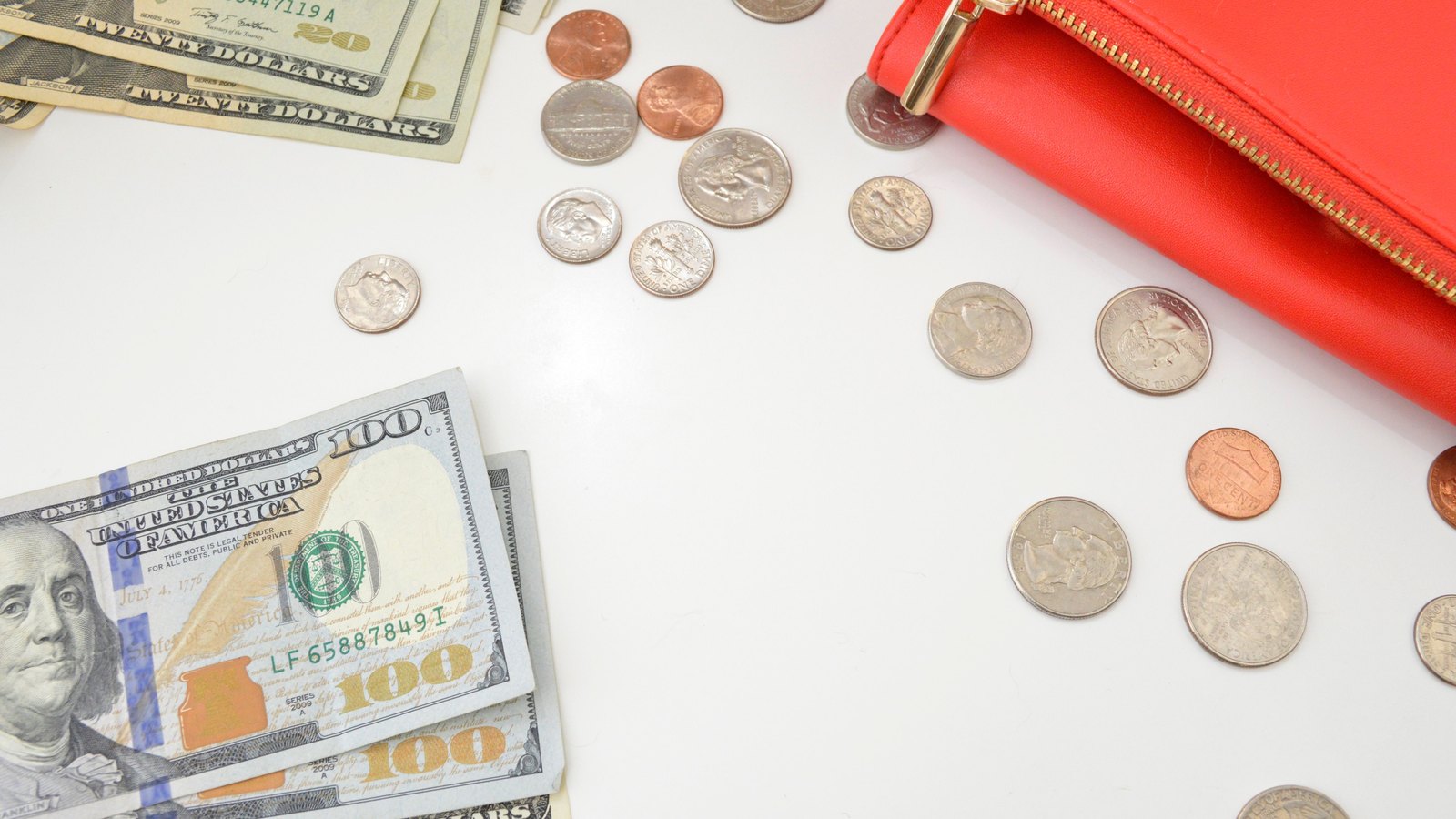 US twenty and hundred dollar bills and scattered coins on a white surface beside an open red wallet.