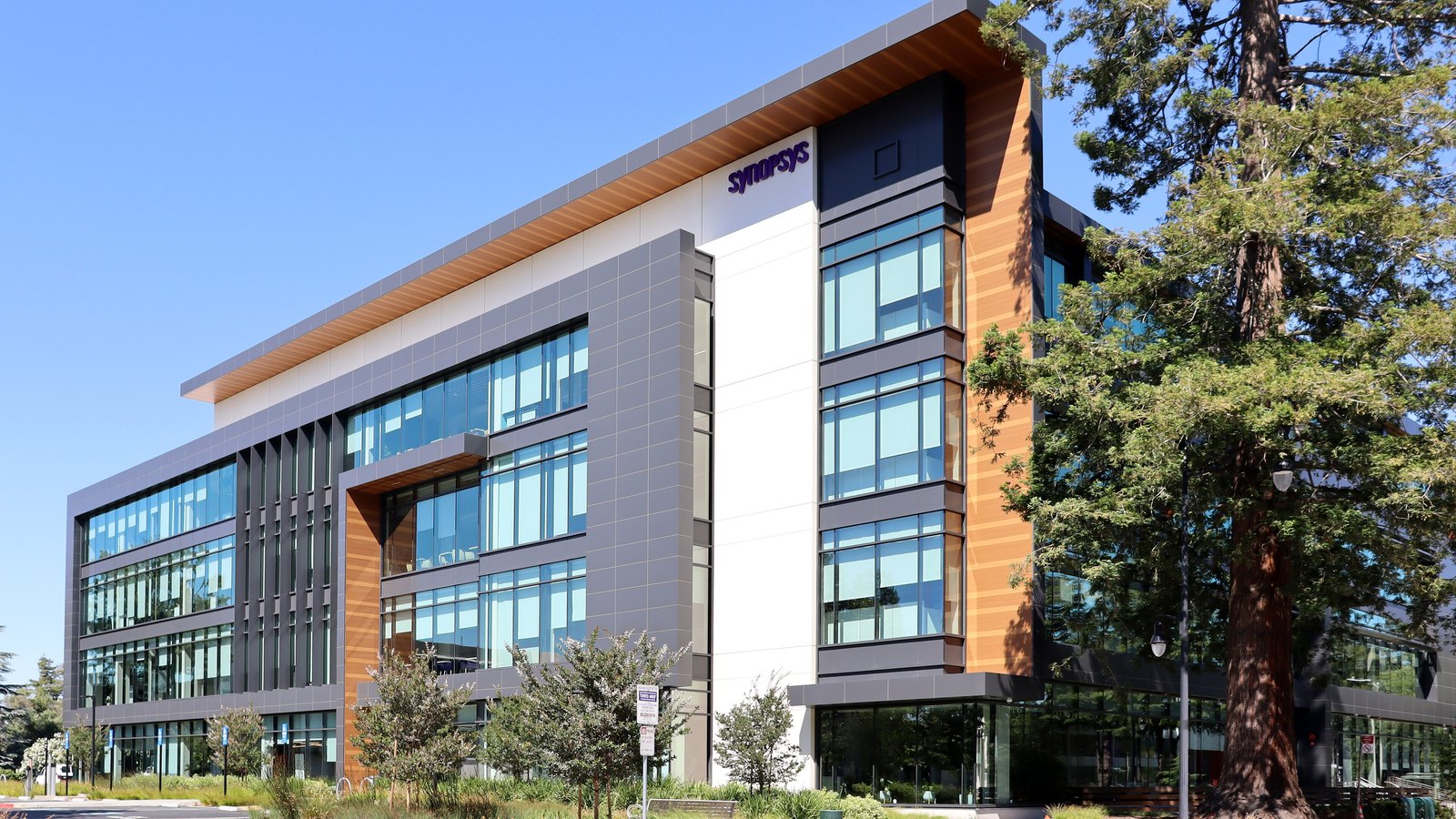 The Synopsys headquarters at 675 Almanor Avenue in Sunnyvale, California, photographed from the street corner: a four storey office block faced in grey panels, white render and vertical timber, with the company wordmark high on the corner bay, young trees and a mature redwood in front of it and a clear blue sky behind.
