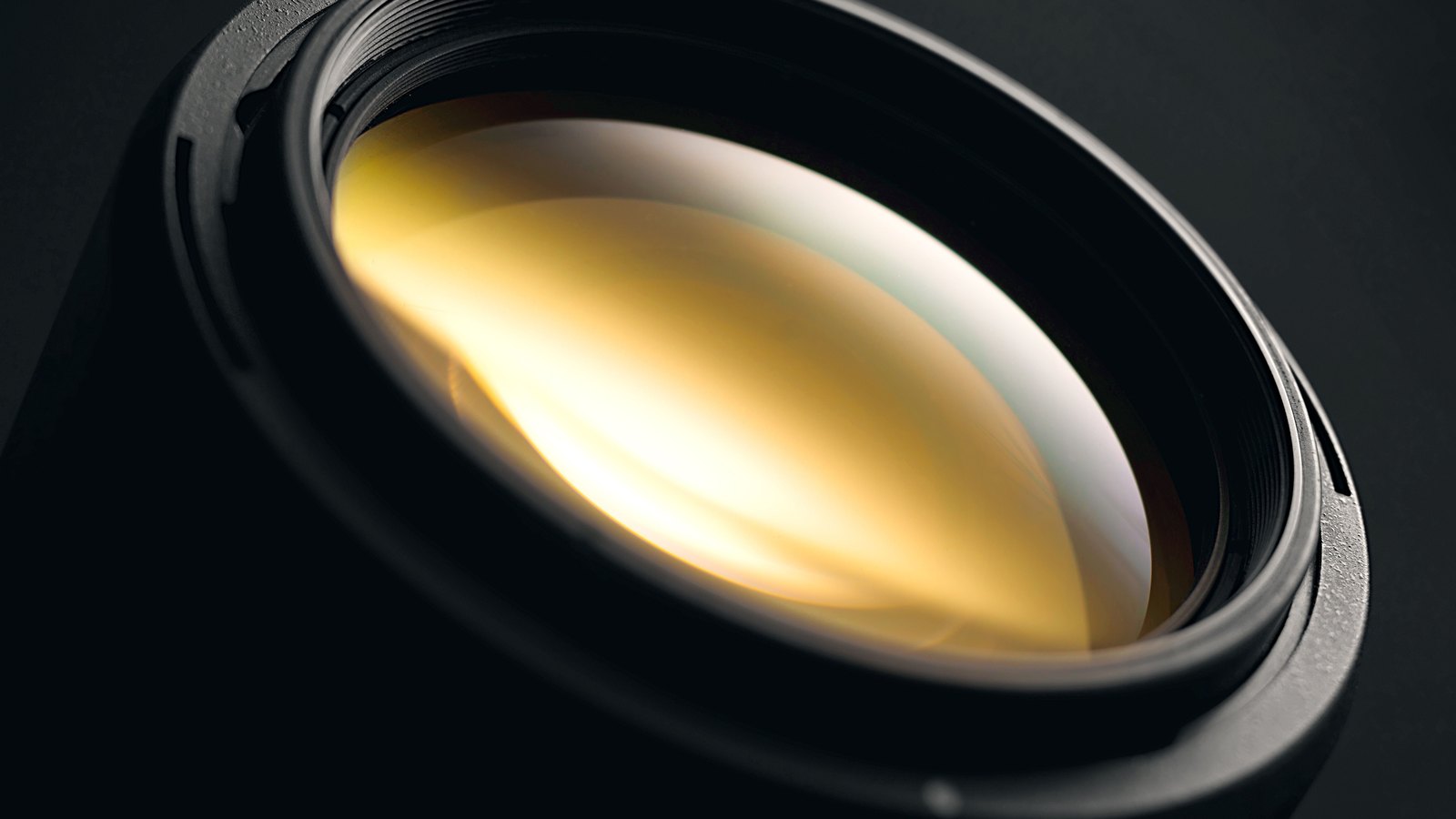 An extreme close up of a coated optical lens element seated in a dark barrel, the curved glass filling the middle of the frame with a warm amber and pale green sheen from its coating, a plain machined metal ring around the rim and an unlit dark grey background. No lettering, marking, brand or person is in view.