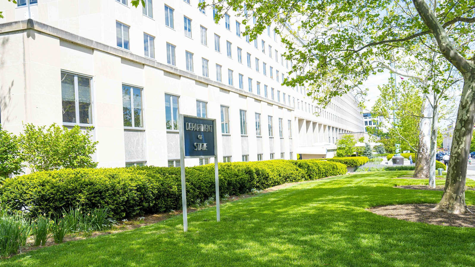 The Harry S. Truman Building in Washington photographed from its lawn in spring, the pale stone facade running away to the right behind clipped hedges and a plane tree, with a plain metal sign on two posts in the foreground reading Department of State.