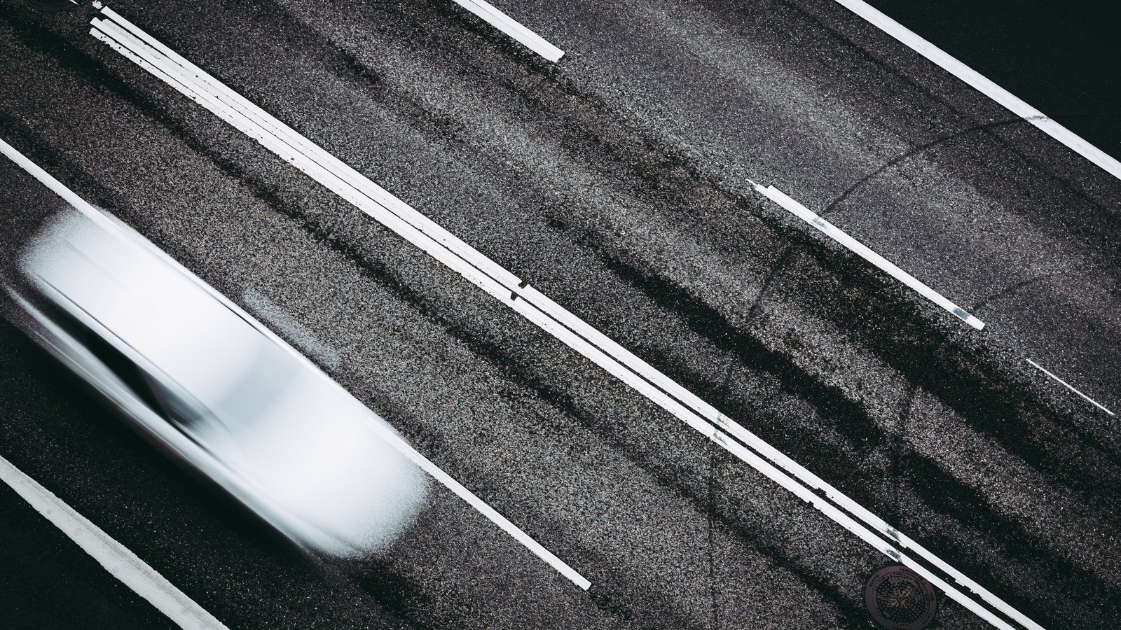 An overhead black and white view of a multi-lane asphalt road with painted lane markings and a single motion-blurred vehicle.