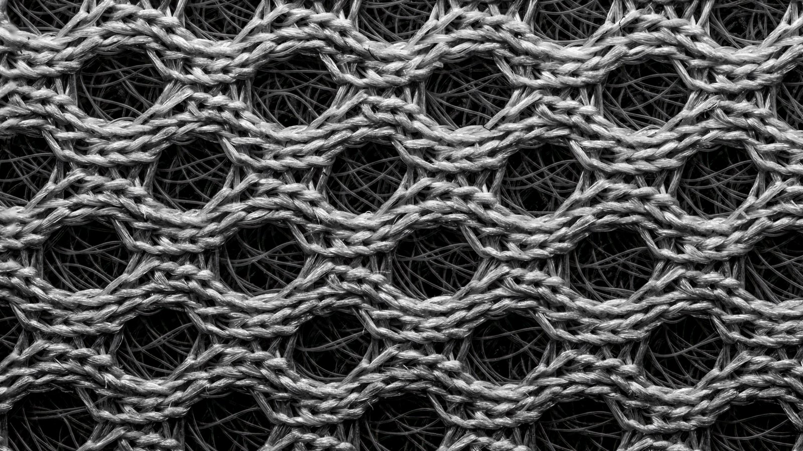 A close macro of open knitted grey mesh, rows of twisted filaments forming an even lattice of oval holes over a darker fibrous backing.