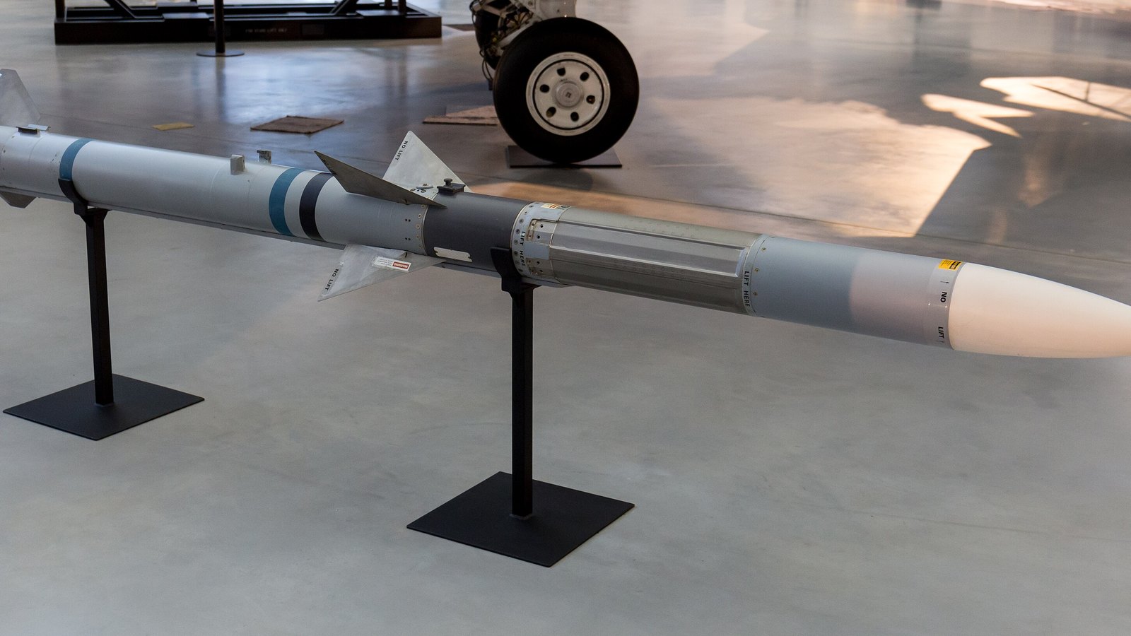 An AIM-120 AMRAAM air to air missile displayed side on across two black metal stands on a bare concrete floor: a pale grey body with blue identification bands, cruciform mid body fins and tail fins, a clear window section behind the mid body and a white nose cone. Stencilled handling instructions reading NO LIFT and LIFT HERE run along the casing. Part of an aircraft main landing gear wheel stands on the floor behind it.