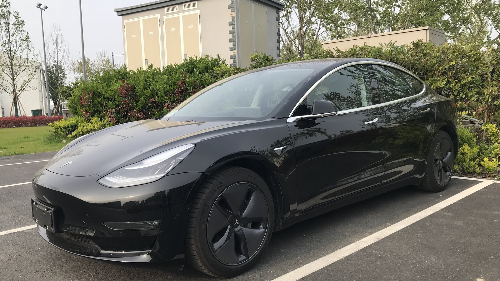 A black Tesla Model 3 parked in a marked bay, seen from the front and side, with the flush recessed door handles visible on both doors and no registration plate fitted to the front bracket.