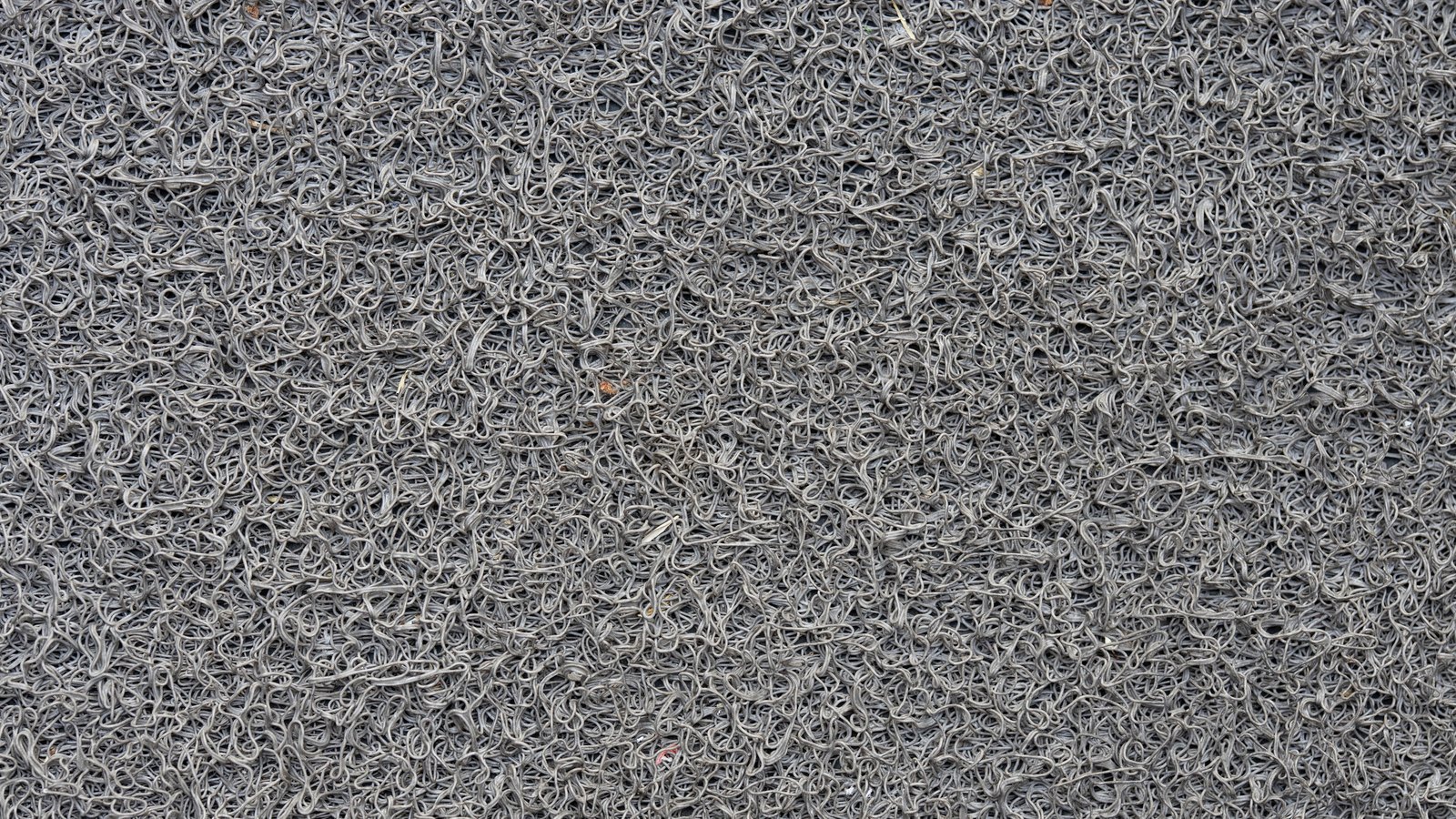 A close overhead photograph of grey looped vinyl matting filling the whole frame, thousands of tangled extruded strands pressed into a dense open mesh, with a few tiny specks of orange and pink debris caught among them. No text, markings, brand marks, people or machinery are in view. A generic moulded polymer close-up, not a photograph of any factory, vehicle or battery.
