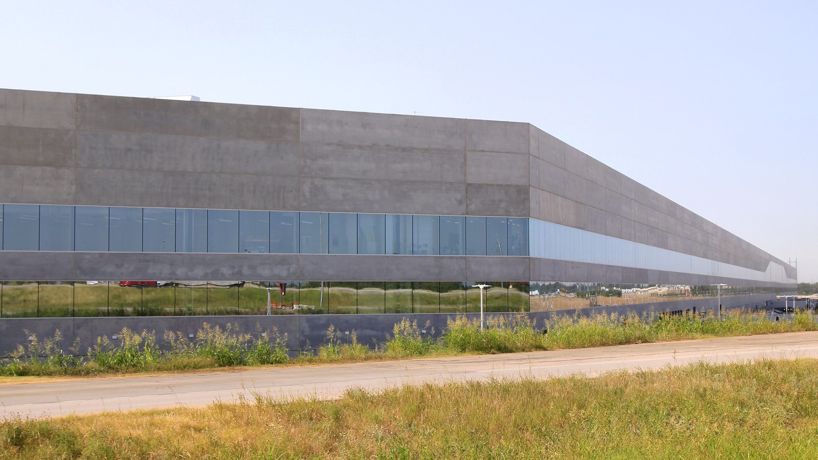 The long grey concrete wall of a factory building with two bands of glass windows reflecting grass, seen across an empty road and a verge of tall summer grass under a pale sky.