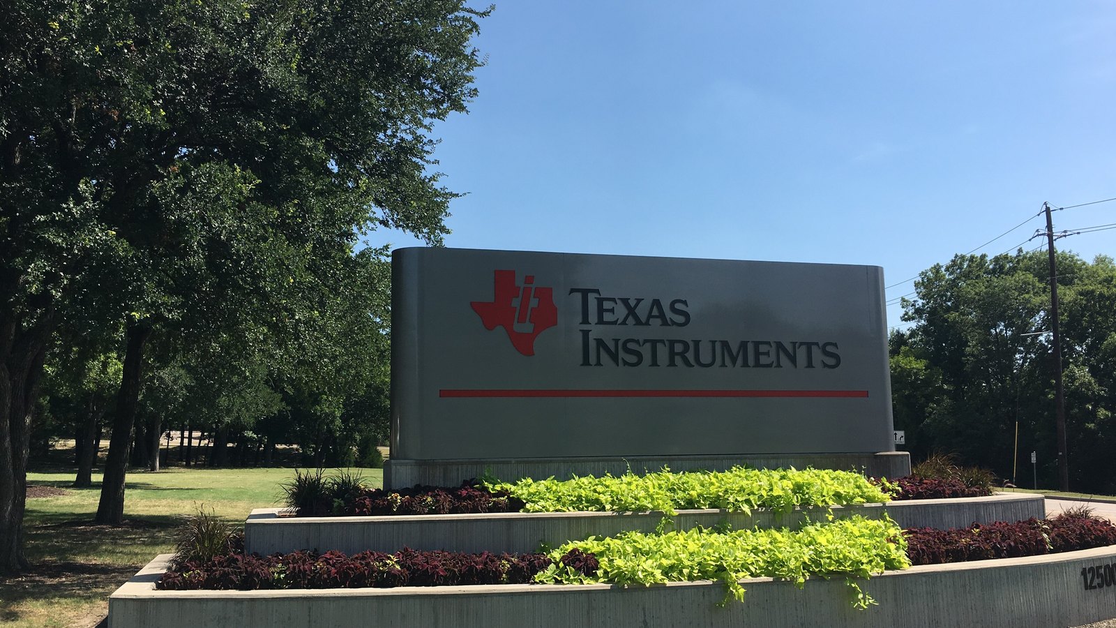 A grey monument sign bearing the Texas Instruments name and logo above tiered flower beds, with oak trees and a clear blue sky behind it.