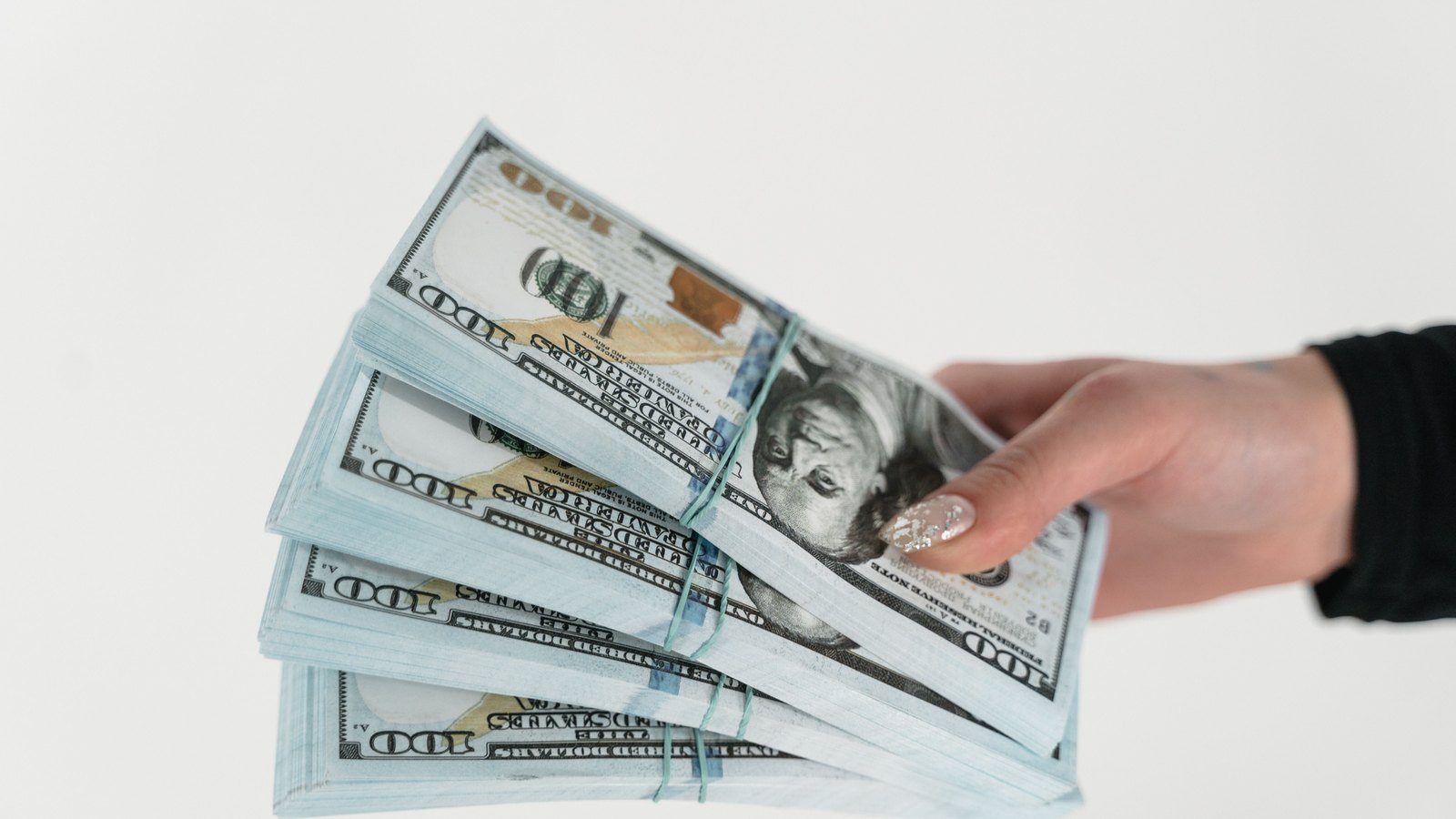A hand holds several thick bundles of US $100 notes against a plain white background.