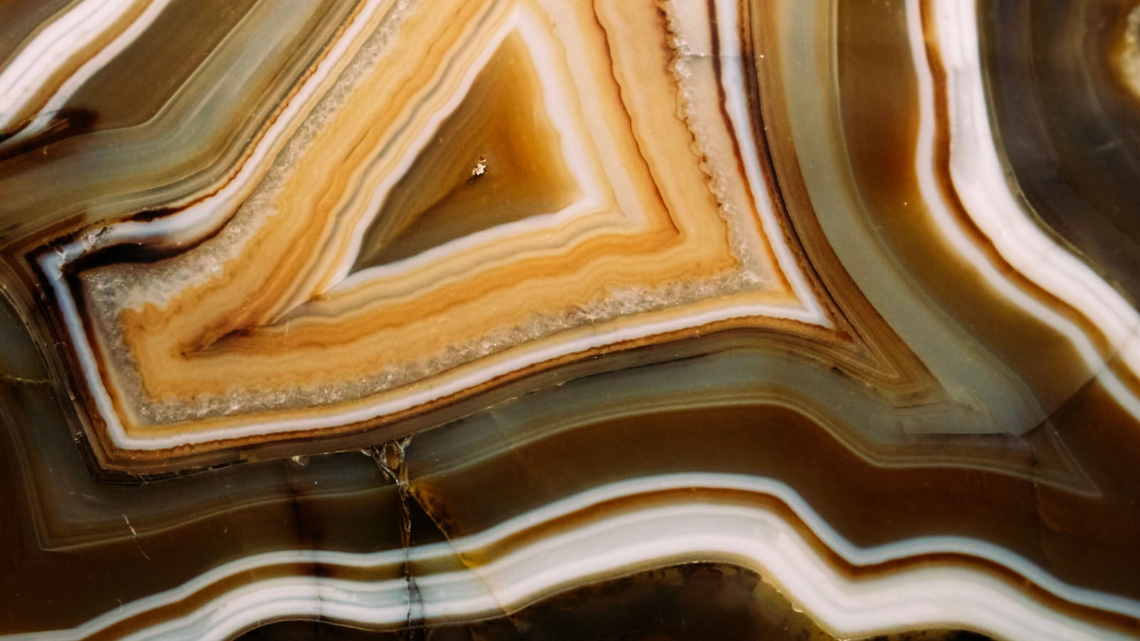 A close photograph of a cut and polished banded agate filling the whole frame. Concentric bands of amber, cream, white and olive brown run as nested angular chevrons across the upper left, and a wider set of parallel wavy bands curves through the lower half. A few fine hairline cracks cross the polished face. No text, markings, brand marks, people or objects are in view. A generic mineral close-up, not a photograph of any document, market or institution.