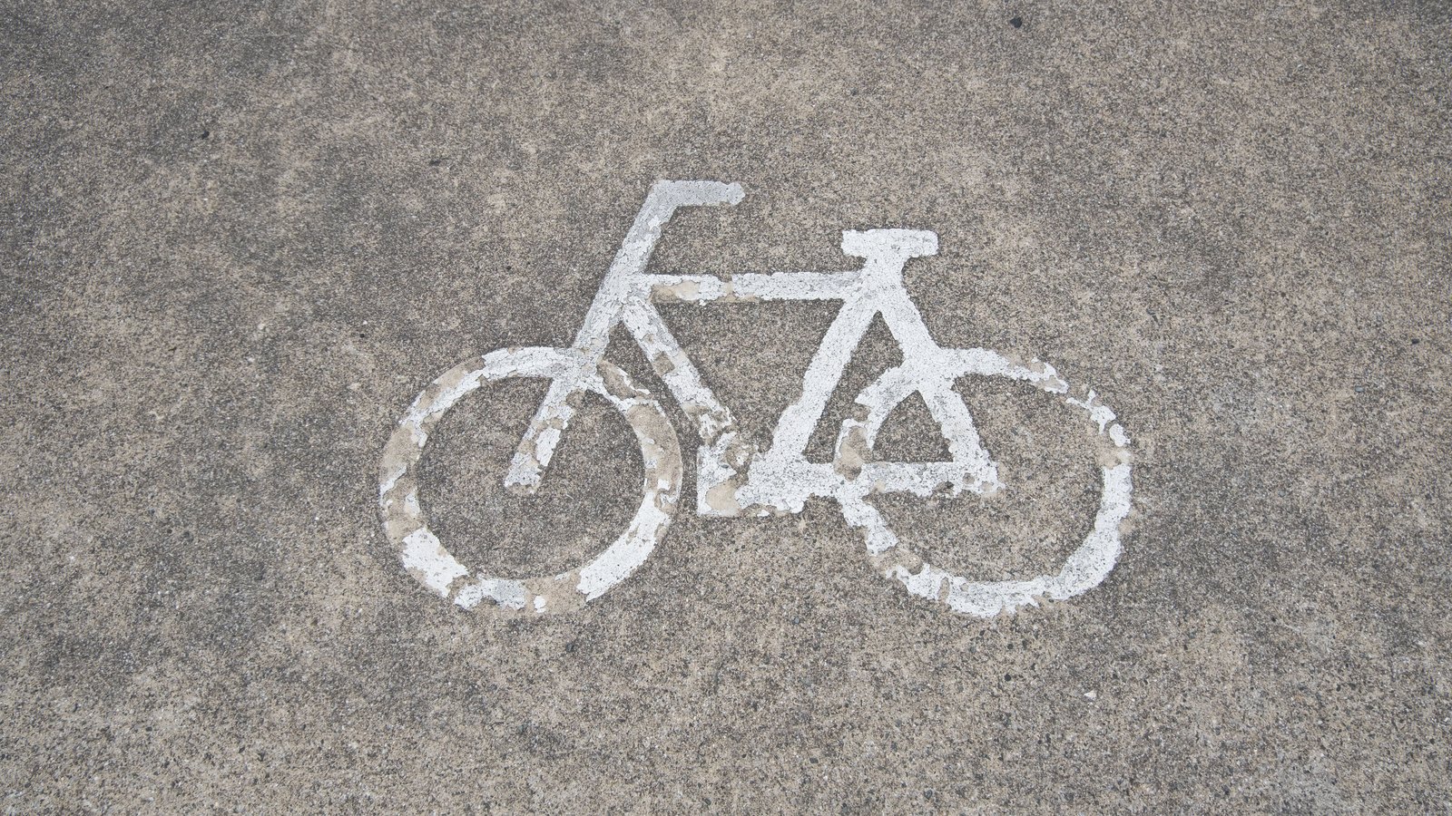 A white bicycle symbol painted on grey pavement, seen from above, with no people, vehicles, signs or legible text in the frame. A generic illustration of a road bike lane, not a photograph of this event.