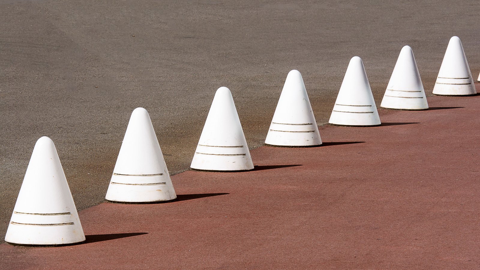 A row of eight white conical concrete bollards, each banded with two thin incised lines, set in a receding diagonal line along the join between grey asphalt and a rust red paved surface, in hard sunlight that throws each cone's shadow to the right. No people, vehicles, road markings, signage, buildings or legible text are in view, and no place can be identified from it. A generic illustration of lane separation on a road surface, not a photograph of Three Oaks Parkway, of Old Fiddlesticks Boulevard or of any Lee County road.