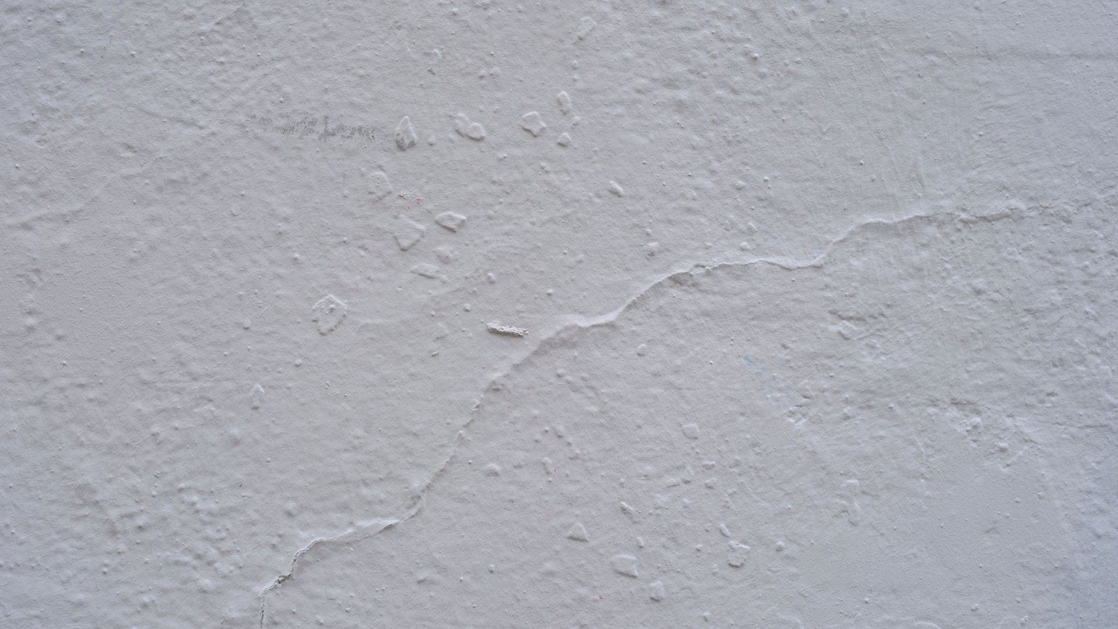 A close crop of a rough off white concrete surface, lit flat, with a shallow hairline crack running across it and small pits and flecks in the render. No street, house, driveway, vehicle, animal, person, text or signage is visible. A generic illustration, not a photograph of Nuna Avenue or of the incident described.